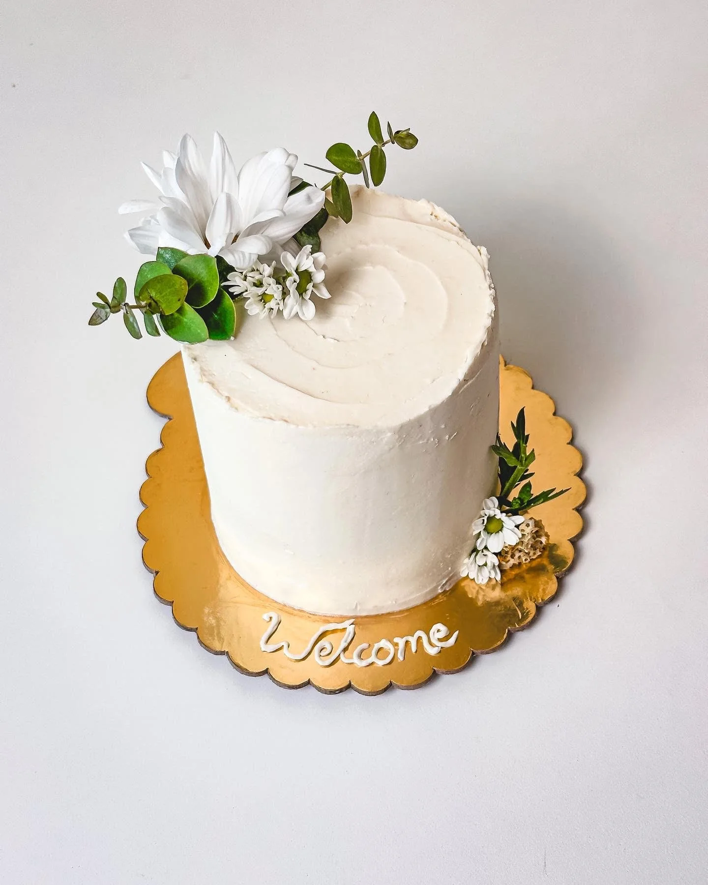 White cake decorated with white flowers and greenery on a golden scalloped cake board with 'welcome' written in white icing.