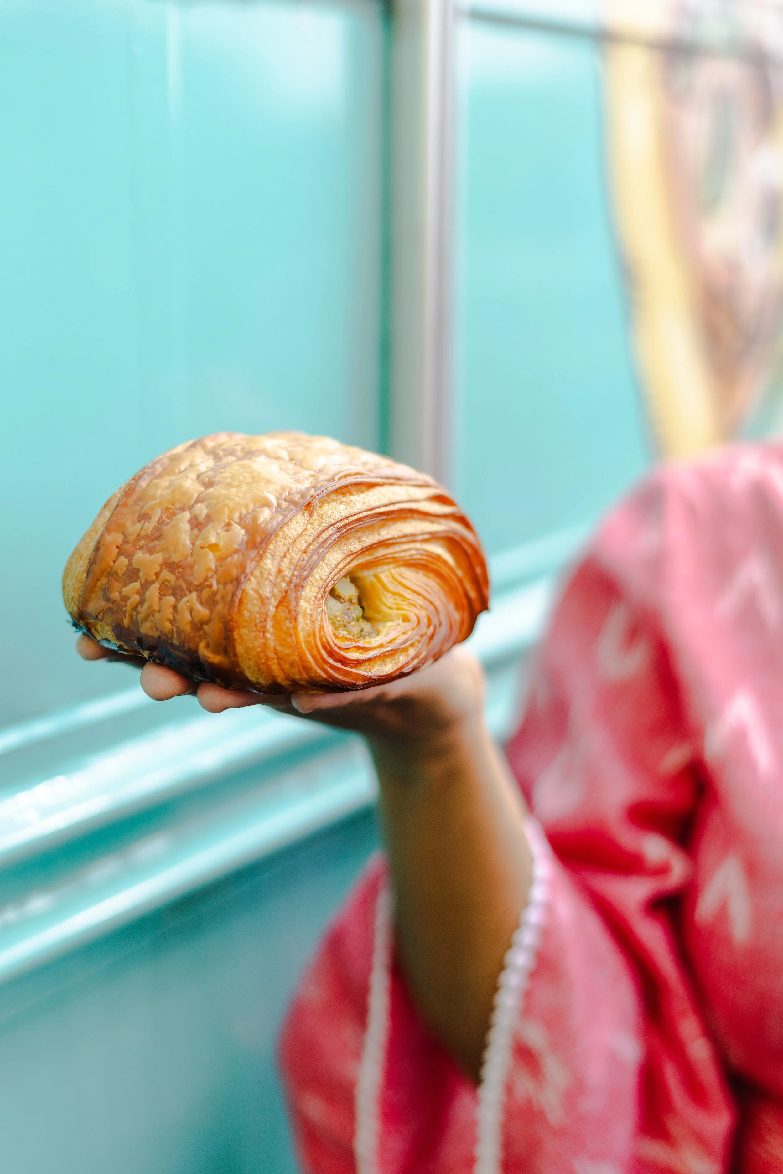 A person dressed in a pink outfit holding a large, flaky croissant with a swirl pattern, against a pastel-colored background.