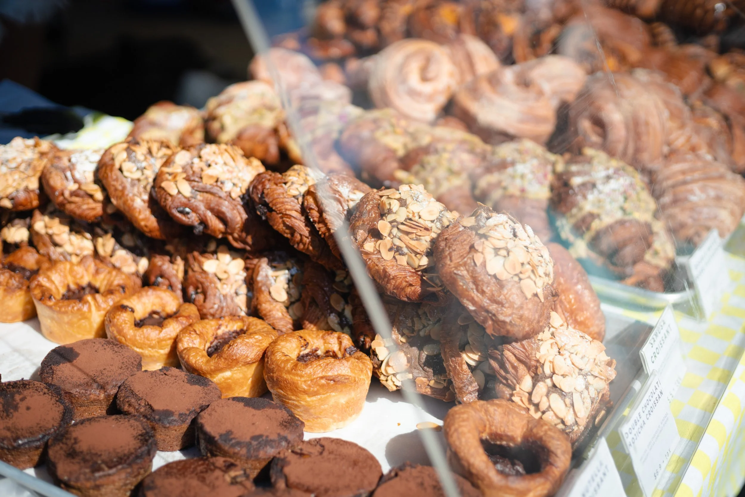 Assorted pastries including croissants, muffins with almond slices, and chocolate treats displayed behind a glass case at a market or bakery.