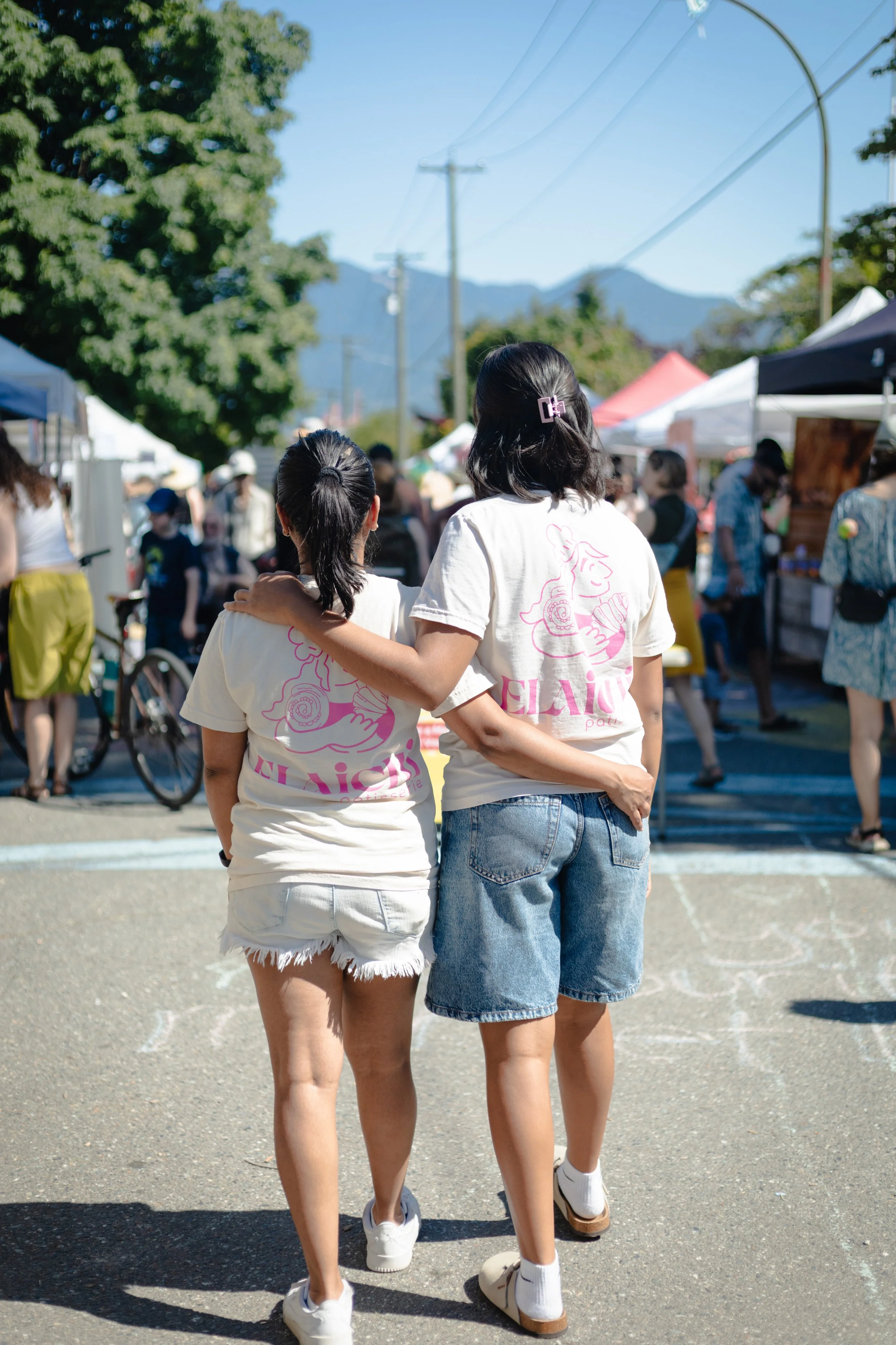 Two people, one wearing white shorts and the other in denim shorts, walk arm-in-arm at an outdoor market wearing Elaichi Patisserie T-Shirts. There are market tents and people in the background, trees, and mountains.