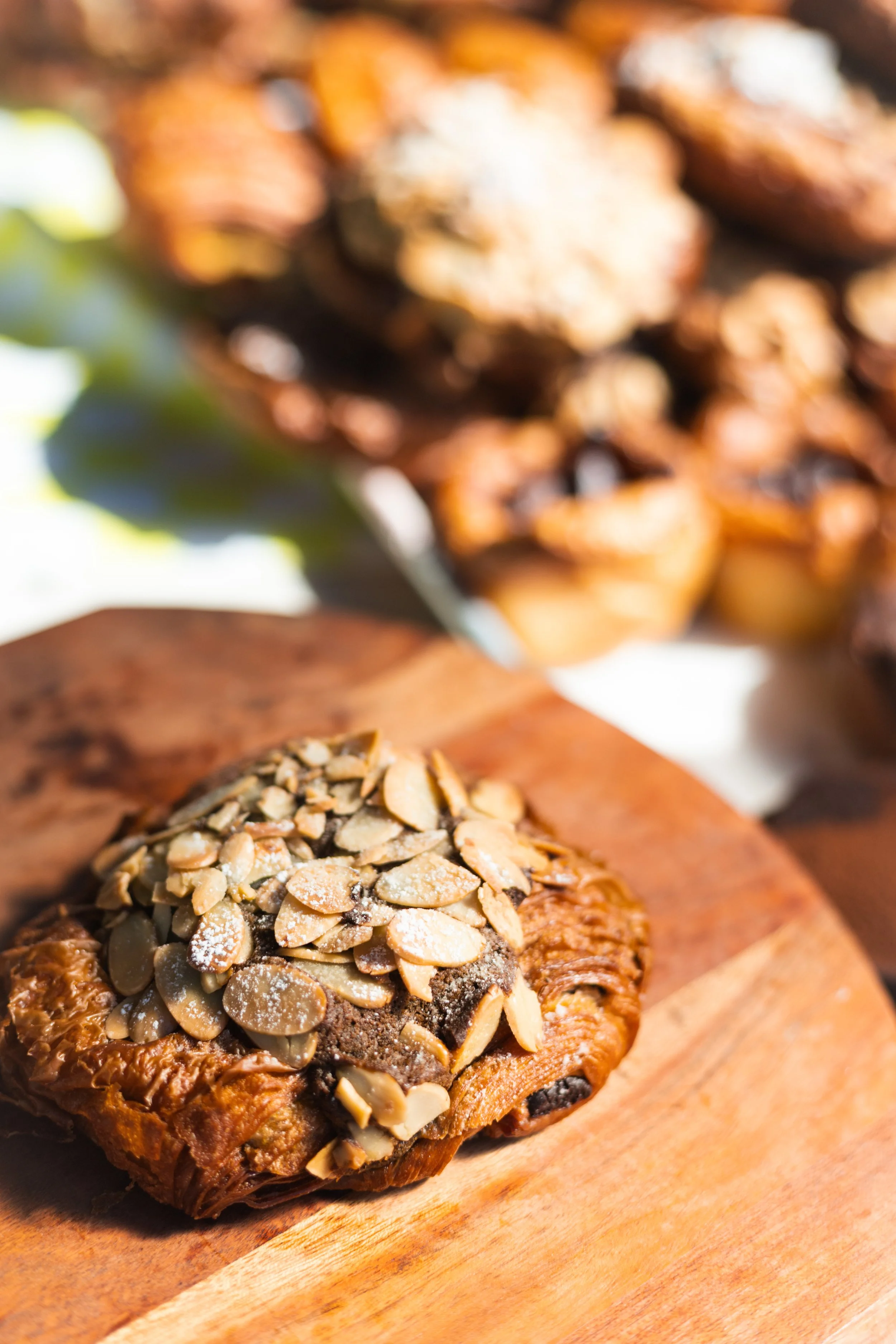 Close-up of a chocolate pastry topped with sliced almonds on a wooden cutting board, with more pastries in the blurred background.