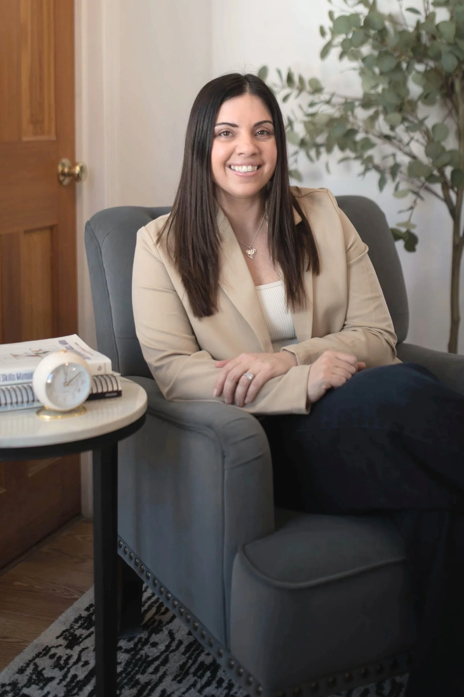 A smiling woman with long dark hair wearing a beige blazer sitting on a gray armchair in a cozy room with a plant in the background. There are books and a small clock on a side table beside her.
