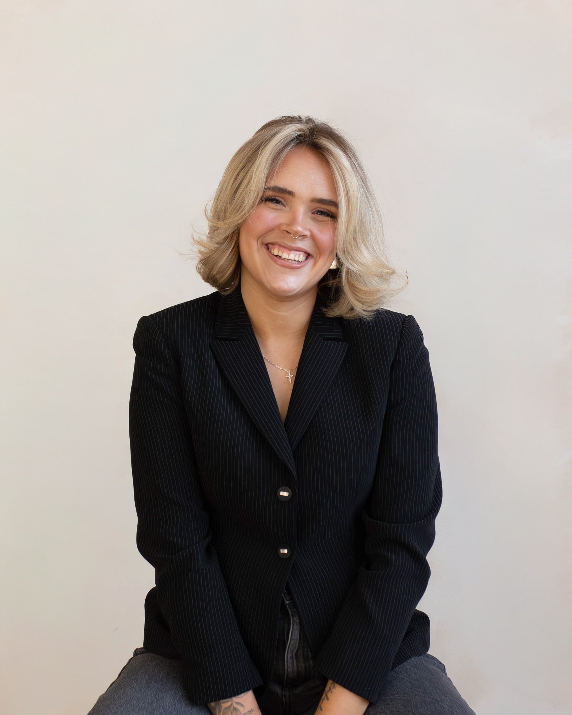A woman with shoulder-length blonde hair, wearing a black pinstripe blazer and gray pants, smiling while sitting against a plain light-colored wall.