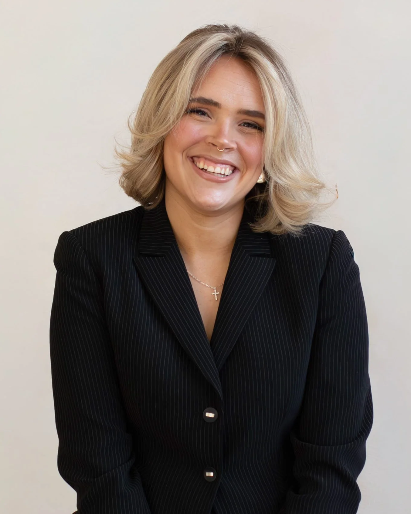A woman with blonde hair wearing a black blazer, smiling, with a necklace featuring a cross pendant, against a plain light background.