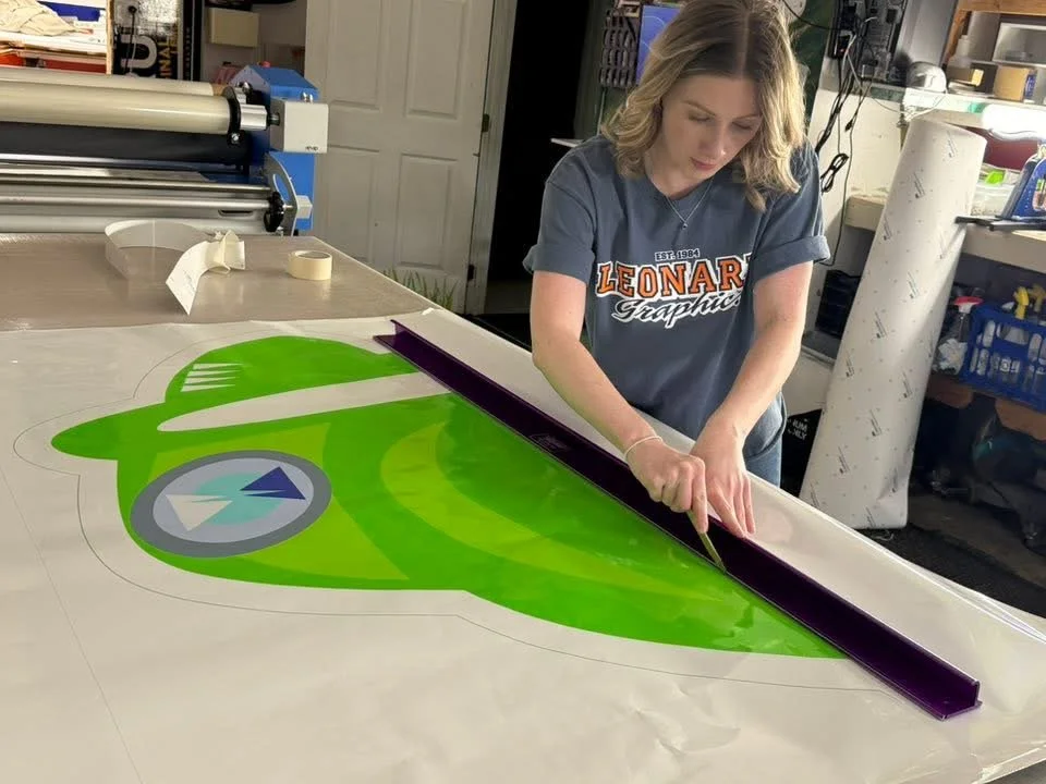 A woman is working on a large green and purple graphic decal in a workshop. She is using a cutting tool on the decal which features a fork and knife design.