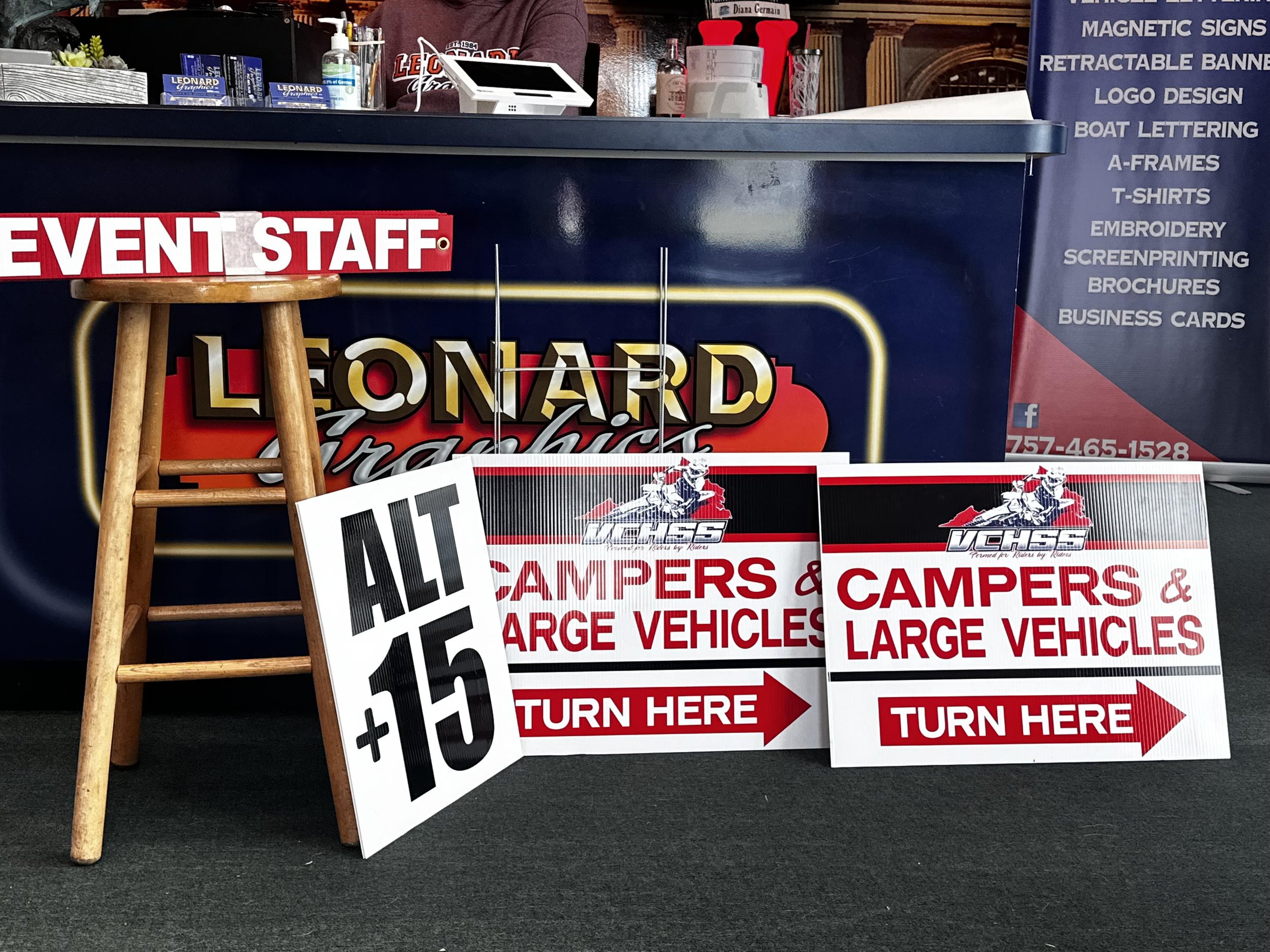 Signage for a vehicle sales event showing signs that read ‘Campers and large vehicles, turn here’ with red arrows, and a separate sign reading ‘Alt 15+’. In the background, a counter with promotional materials, a hand sanitizer bottle, and a person i