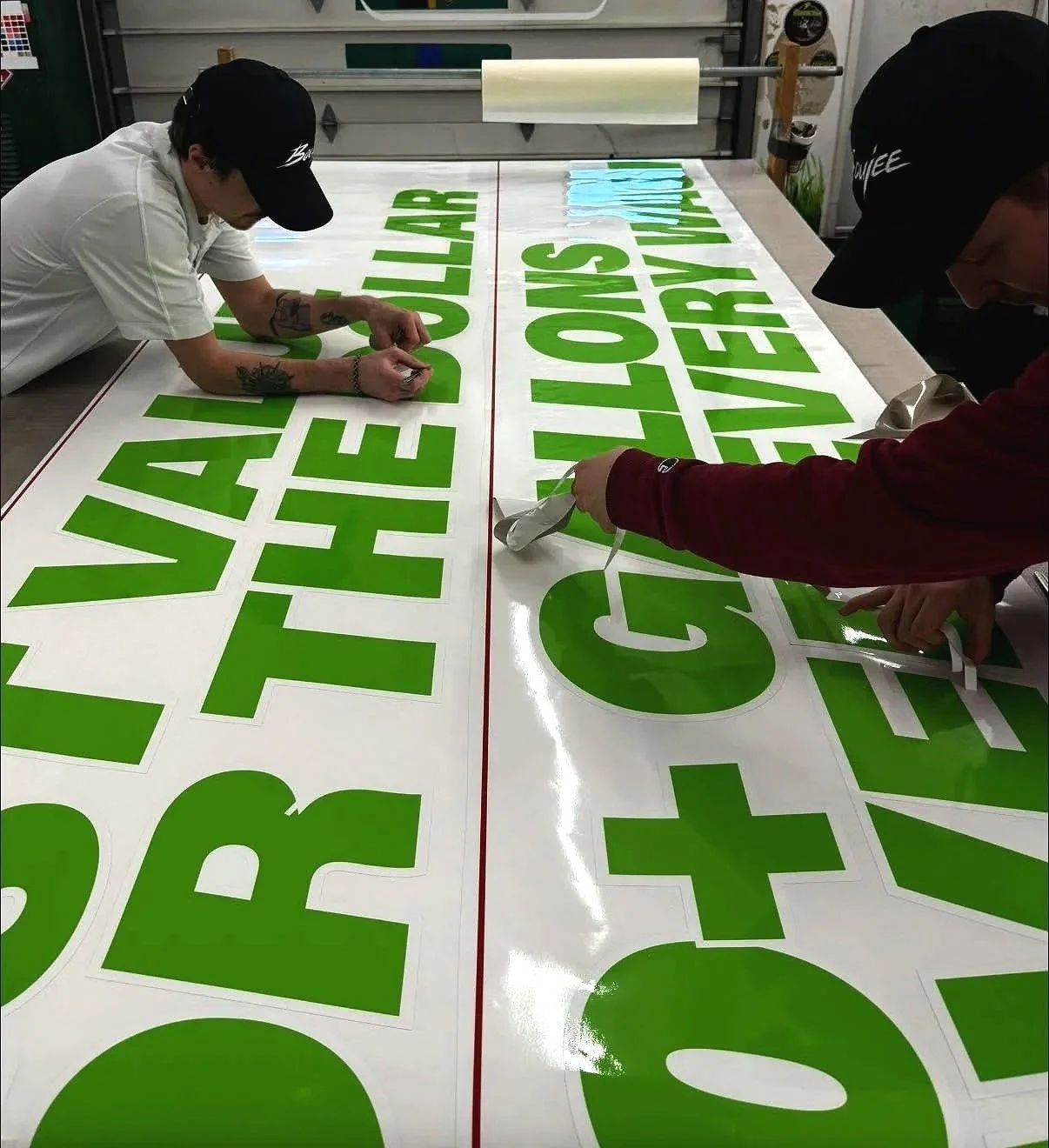 Two individuals are working on applying large green letters to a white banner with red lines for a sign or display.