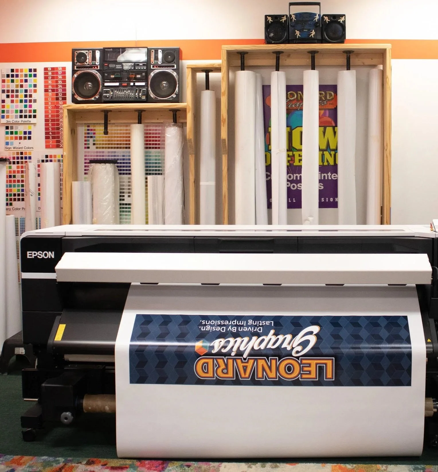 Large Epson UV flatbed printer printing a banner in a sign shop with color swatches and rolled printing materials on shelves in the background.