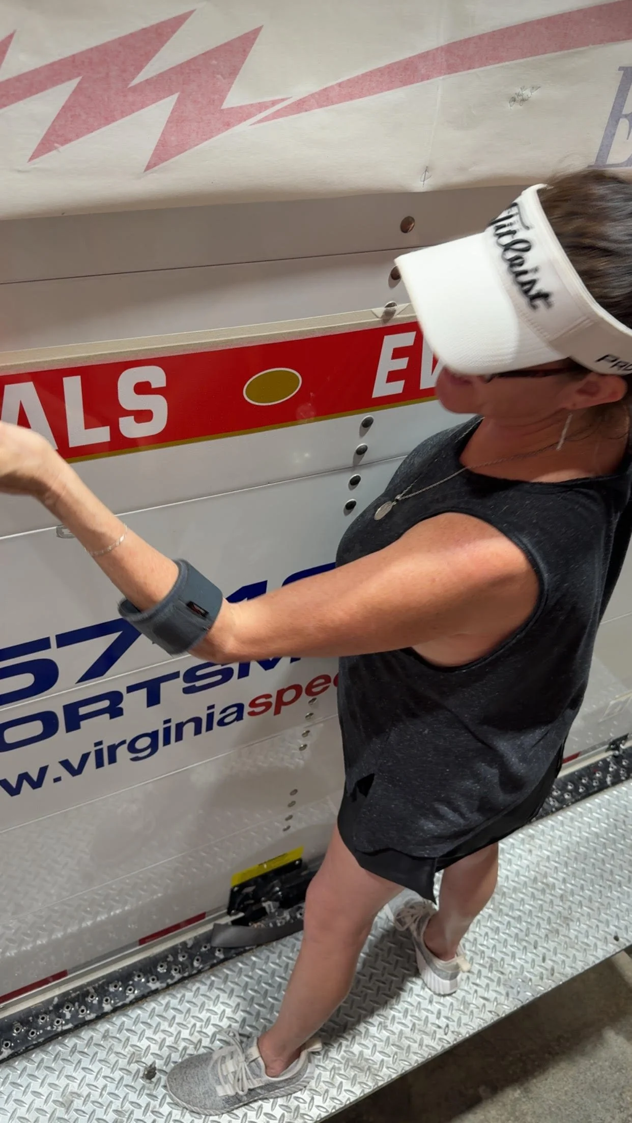 A woman wearing a white visor, black sleeveless shirt, and gray sneakers standing on a metal surface, reaching out to a white vehicle with Virginia Sports Management and Speed Tour graphics.