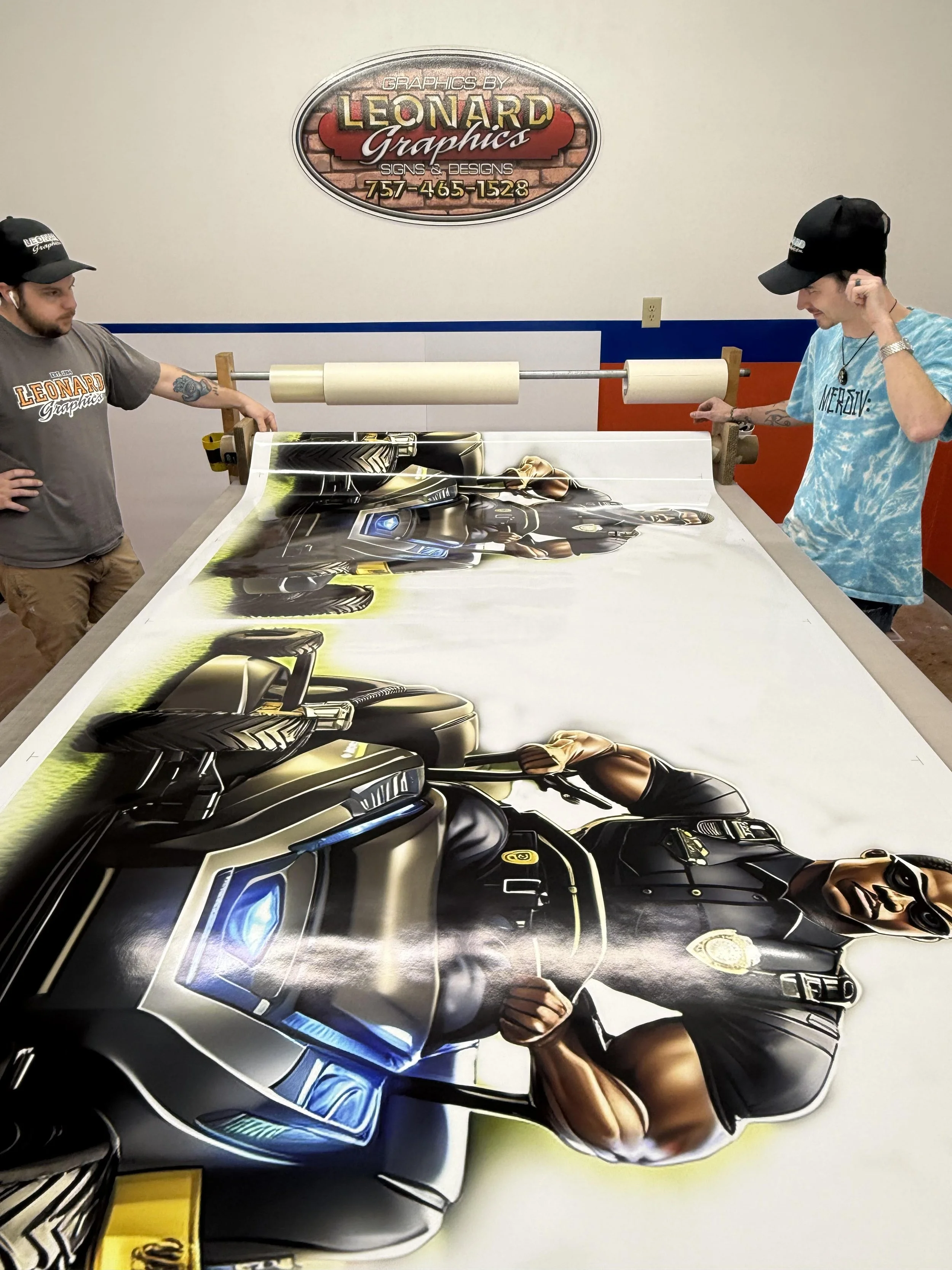 Two men working on a large custom graphic wrap for a vehicle in a sign shop, with a branded sign for Leonard Graphics in the background.