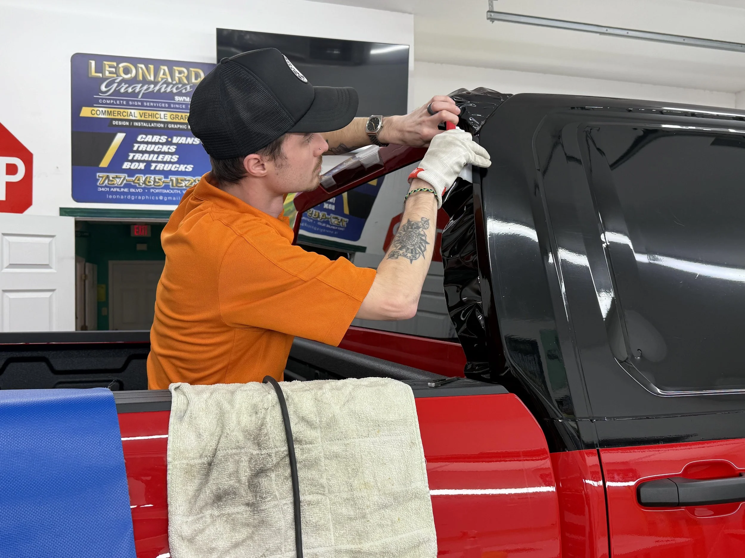 A man in an orange shirt and black baseball cap applies black vinyl film to a vehicle's rear window in a workshop.