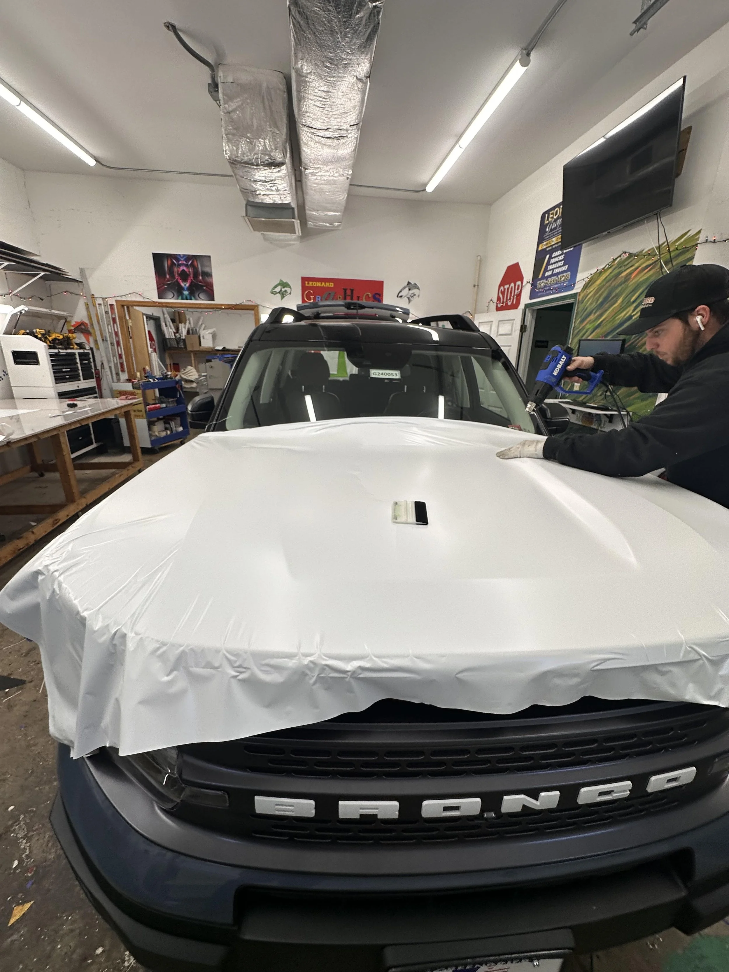 A person working on a black Ford Bronco in a workshop, applying a white protective film or wrap to the hood of the vehicle.