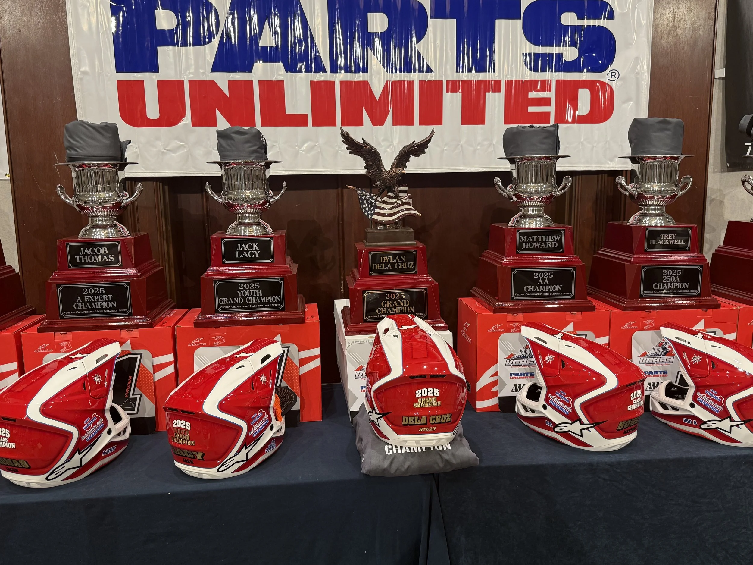 Race helmets and trophies on display with a banner reading "Parts Unlimited" in the background. The helmets are red and white with racing stickers, and the trophies are silver with black bases, each labeled with names and titles, including Jack Thoma