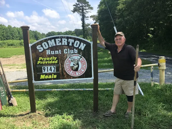 Man holding a fishing rod and a walking stick standing next to a sign for Somerton Hunt Club on a grassy area with trees and blue sky in the background.