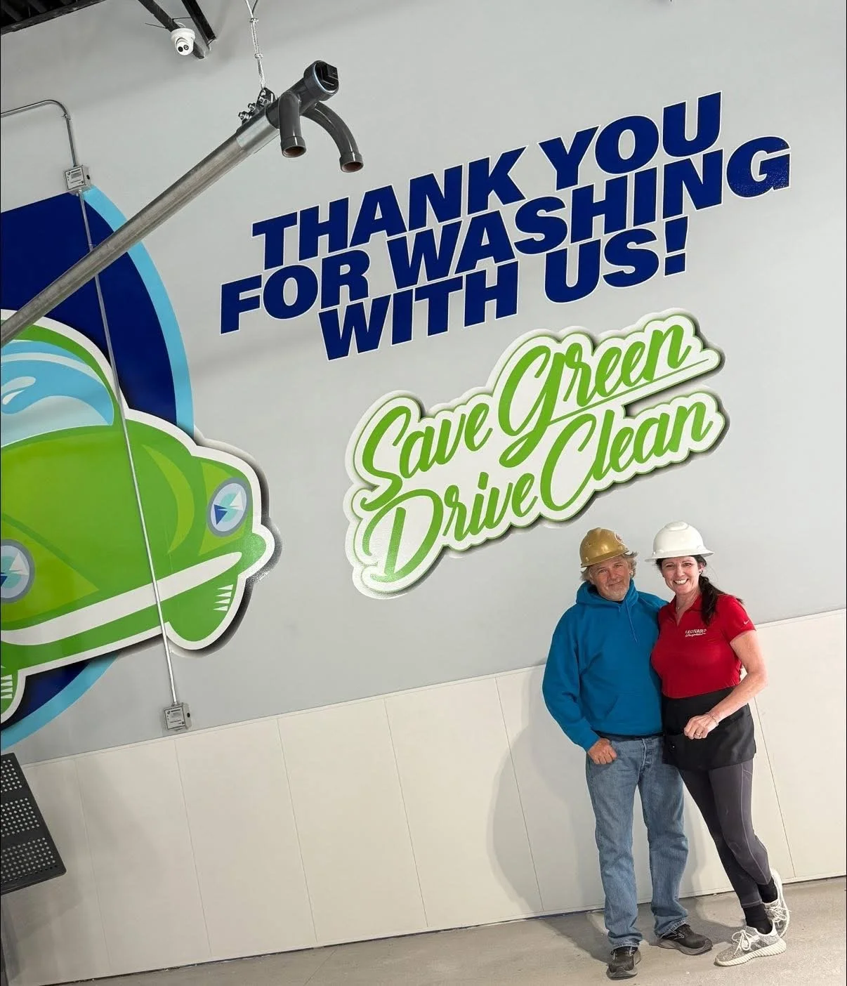 Two people wearing safety helmets standing in front of a sign that says, 'Thank you for washing with us! Save Green Drive Clean,' with a graphic of a green car and a spray nozzle.