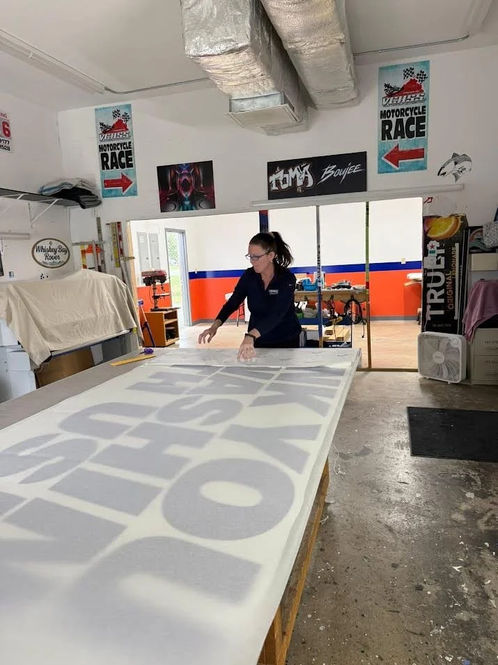 A woman working on a large sign or banner with gray and white lettering in a workshop. posters and signs related to motorcycle racing are visible on the walls, and the space has a workbench and supplies.