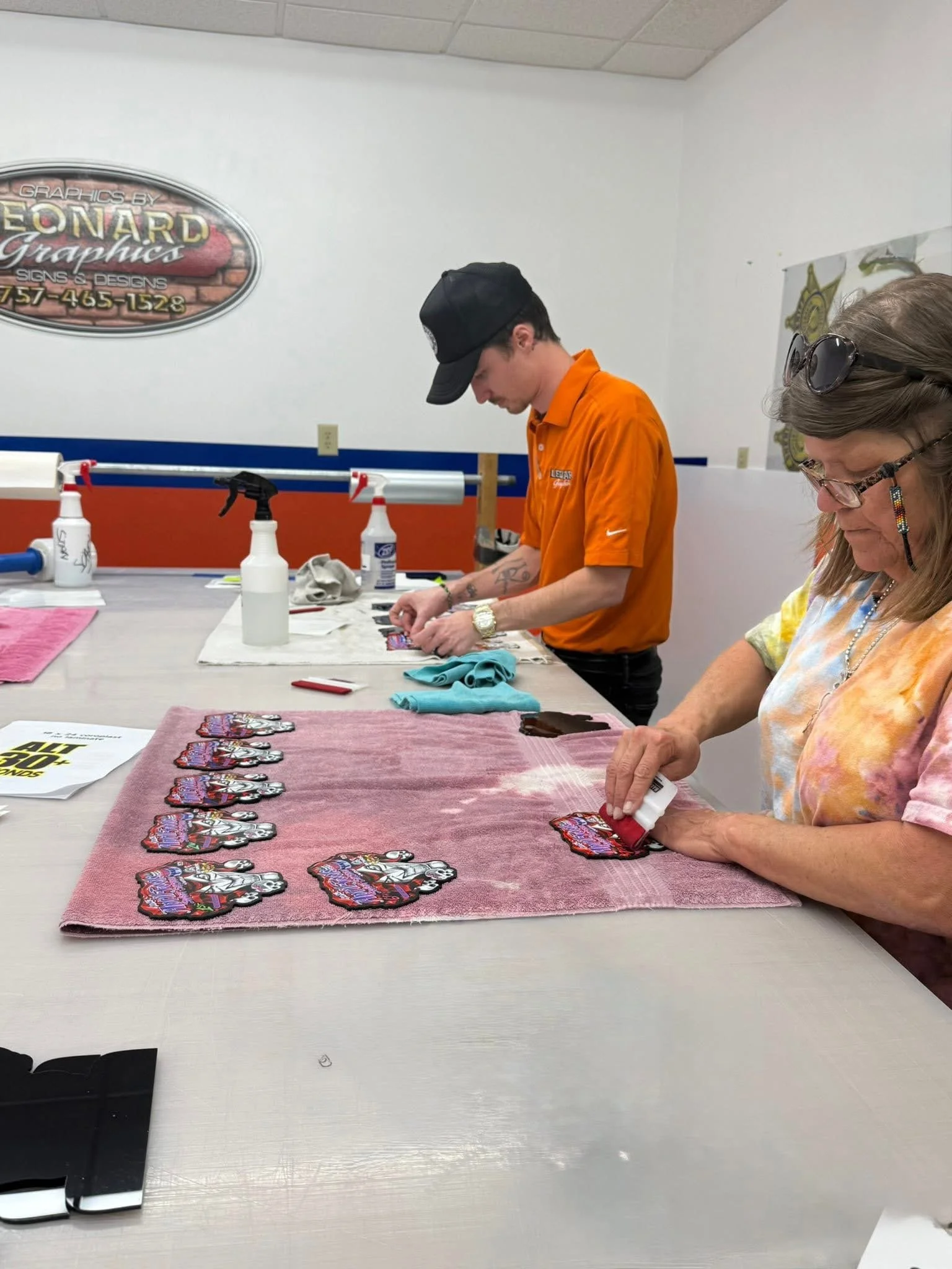 Two people working on custom patches at a table, with approximately ten patches aligned in a row, a pink towel, and various cleaning supplies.