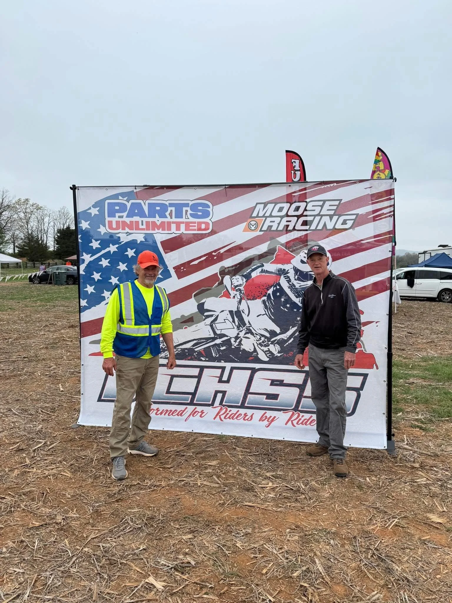 Two men standing in front of a large race banner at an outdoor event. One man is wearing a bright yellow safety vest and an orange cap, and the other man is in a black jacket and gray pants. The banner features the logos 'Parts Unlimited' and 'Moose 