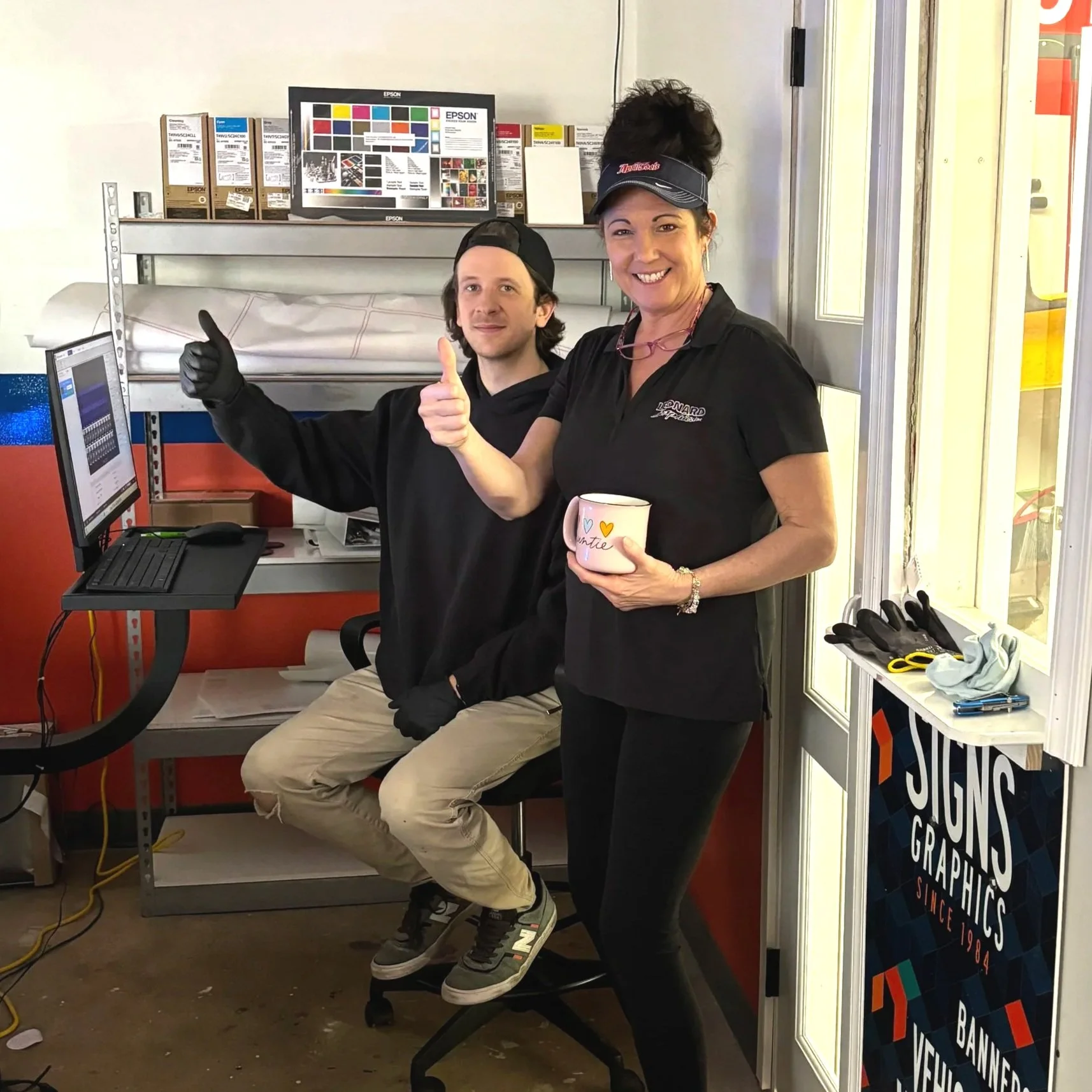 Two smiling people giving thumbs up inside a sign shop, one sitting at a computer and the other holding a mug, surrounded by signage and printing supplies.
