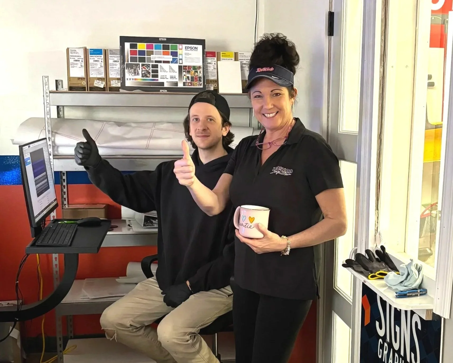 Two women in a sign shop, smiling and giving thumbs-up. One is seated at a computer, while the other is standing and holding a mugs with a handwritten message.