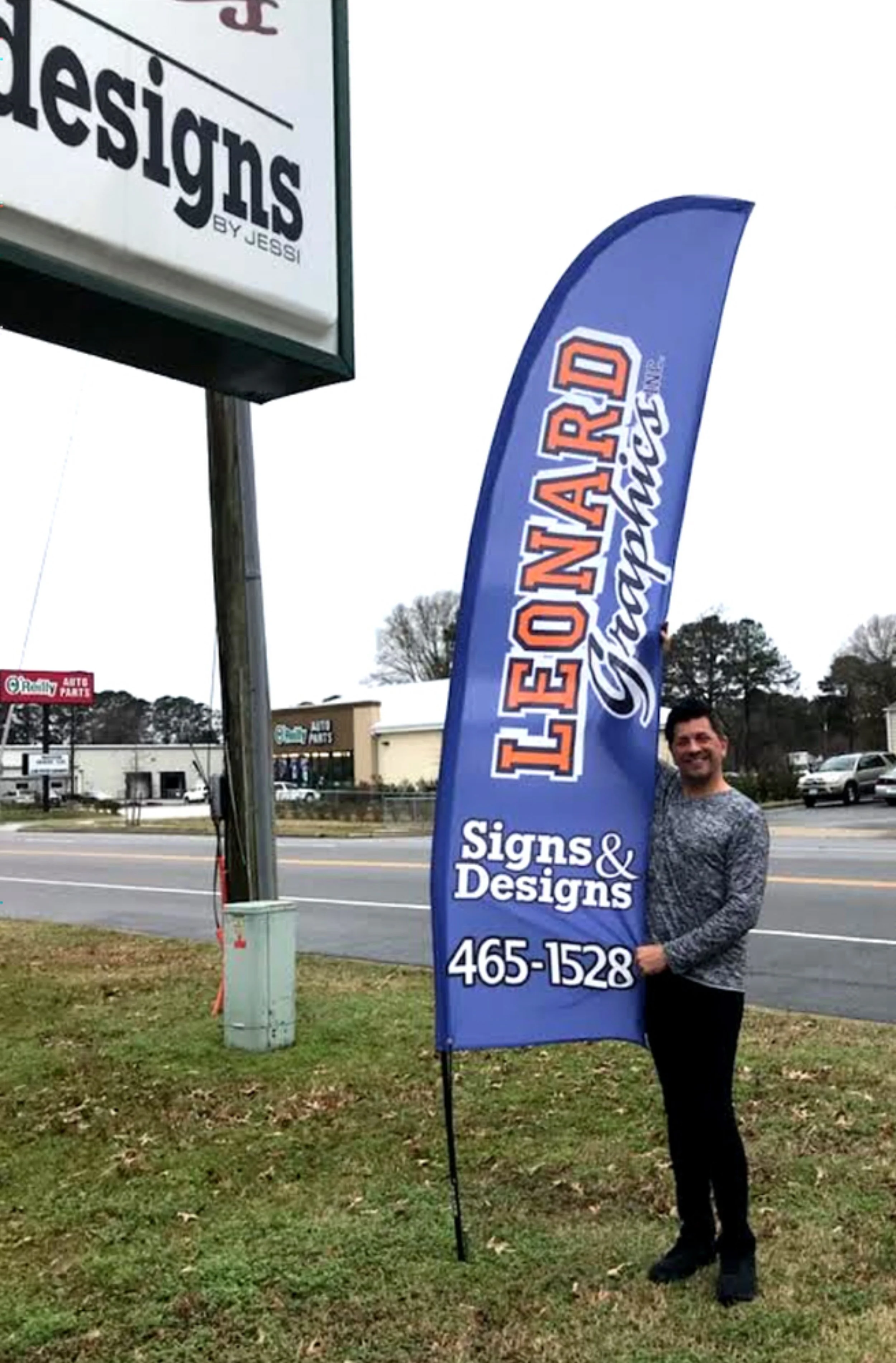 A man standing outdoors on grassy ground next to a tall, blue, feather-shaped flag advertising Leonards Signs & Designs with a phone number. In the background, there is a busy street with cars and buildings, including an auto parts store.