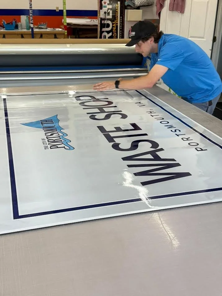 A person in a blue shirt and black cap is applying a large printed sign or banner with text that reads "WASTE SHOOT" and a logo on a flat surface in a workspace.