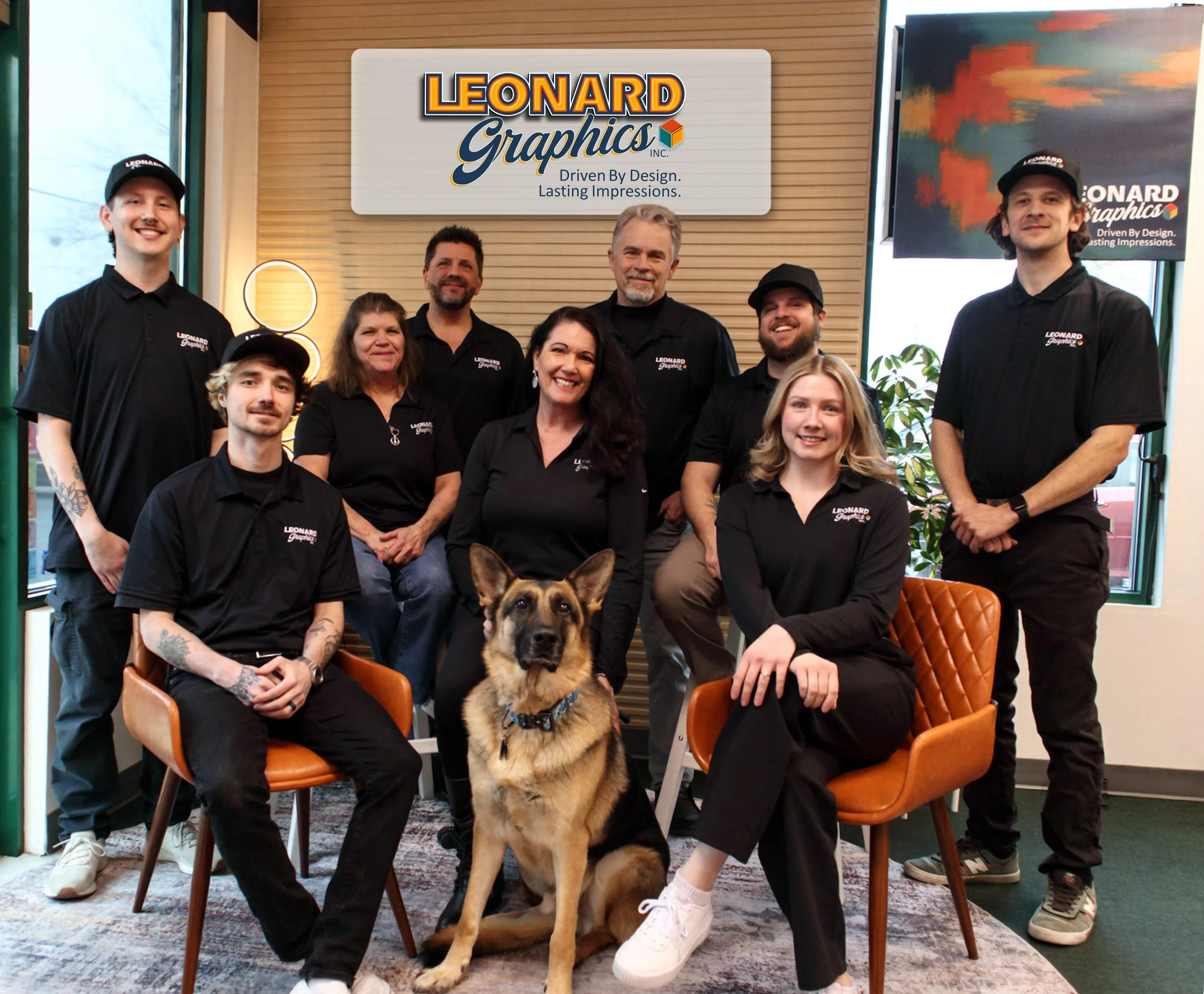 Group of nine people and a dog posing inside Leonard Graphics office, with a sign and a screen displaying the company logo in the background.