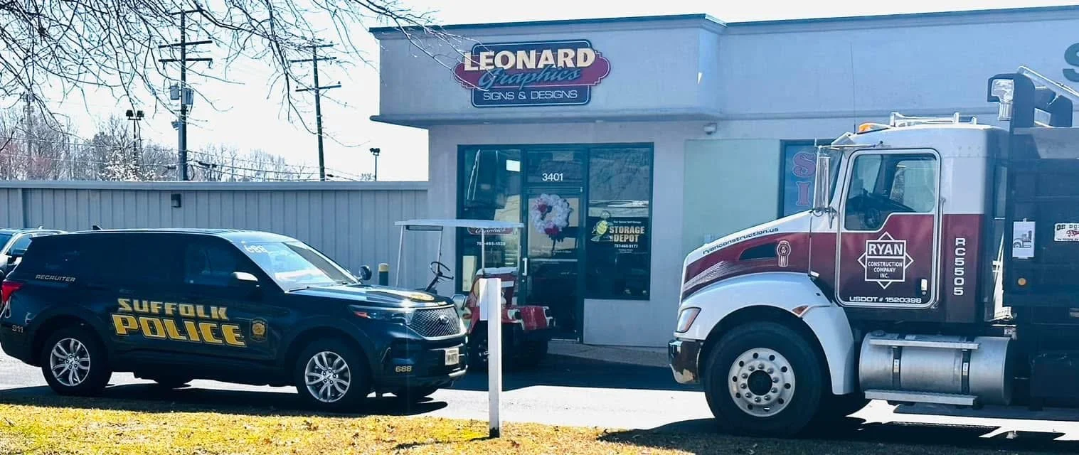 Suffolk Police vehicle and construction truck parked outside Leonard Graphics Signs & Designs storefront.