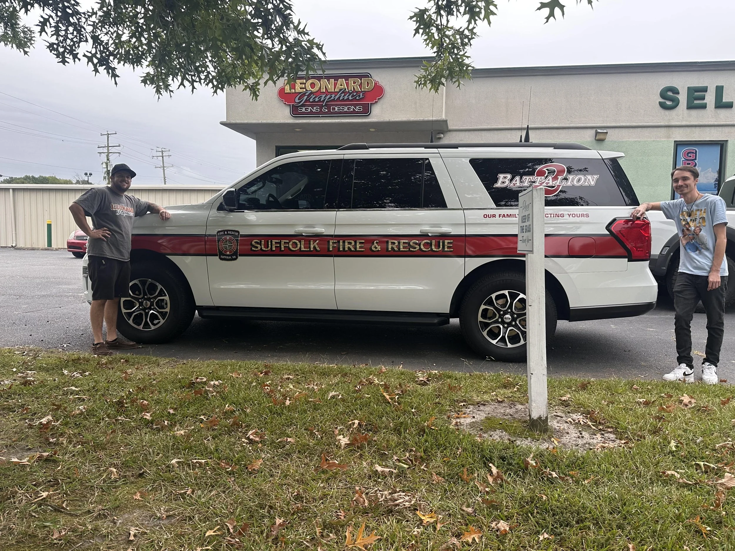 Two men standing beside a Suffolk Fire & Rescue vehicle, which is parked in front of a building with signs for Leonard Graphics and a salon. The man on the left leans on the front of the vehicle wearing a grey T-shirt and shorts, while the man on the