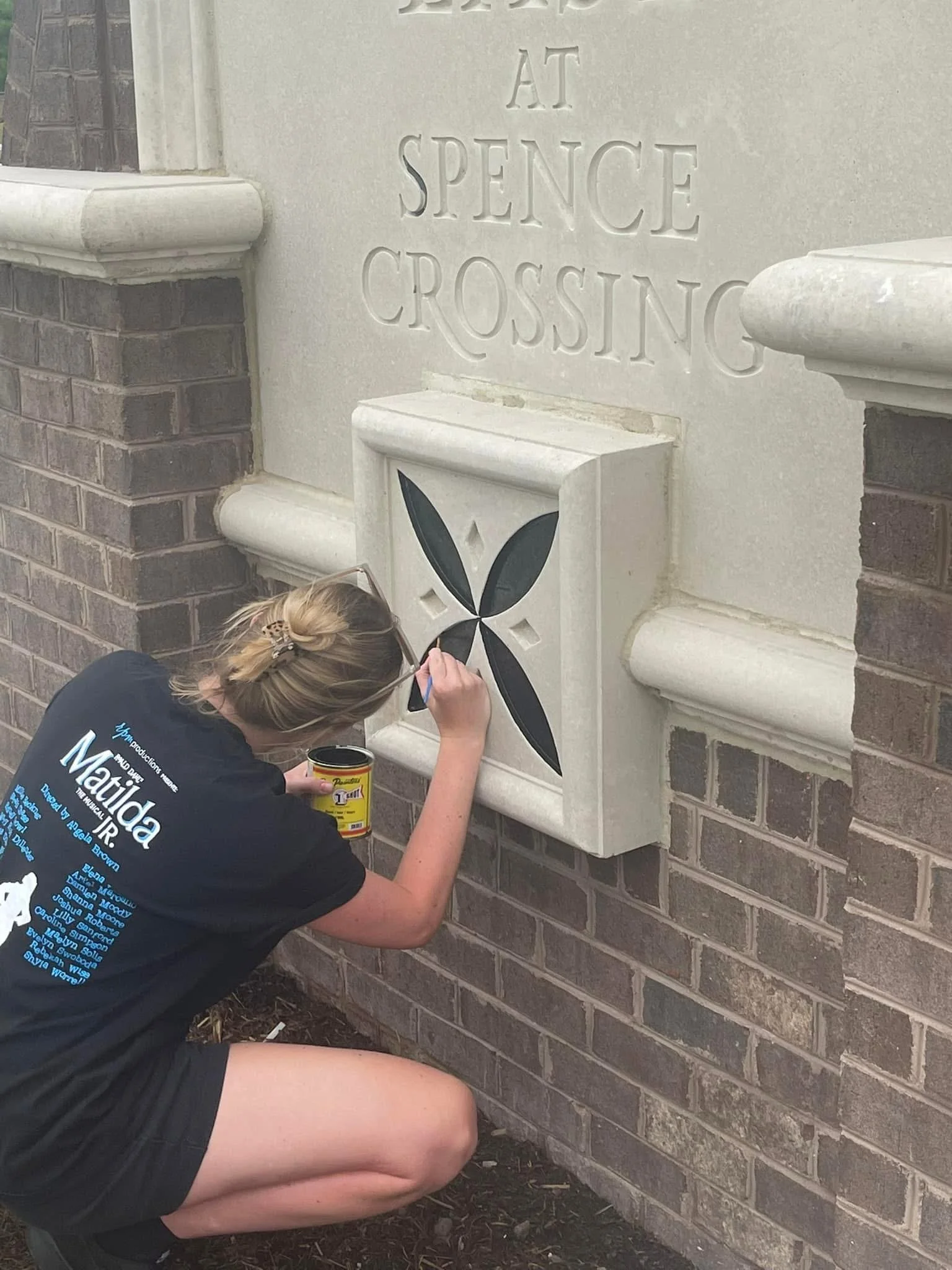 Person painting a decorative design on a brick wall near a stone monument with inscriptions.
