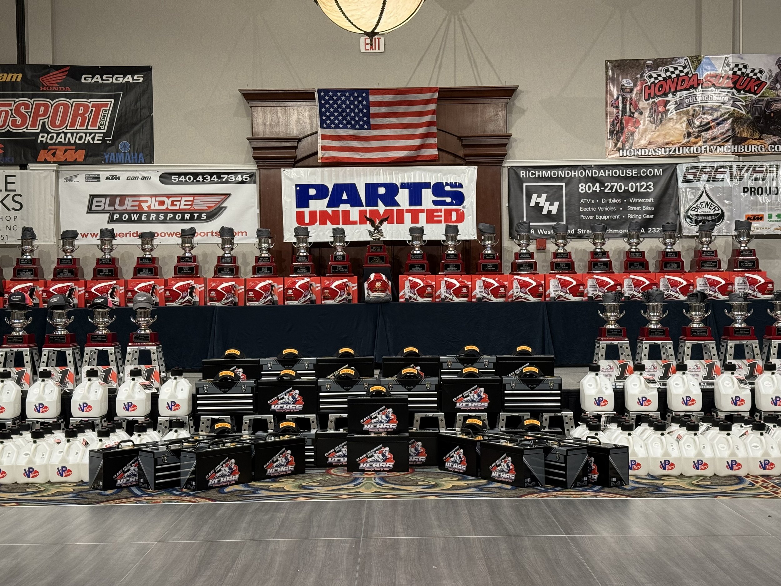 Display of multiple racing trophies, helmets, fuel containers, and gear on a table with banners and flags in the background.