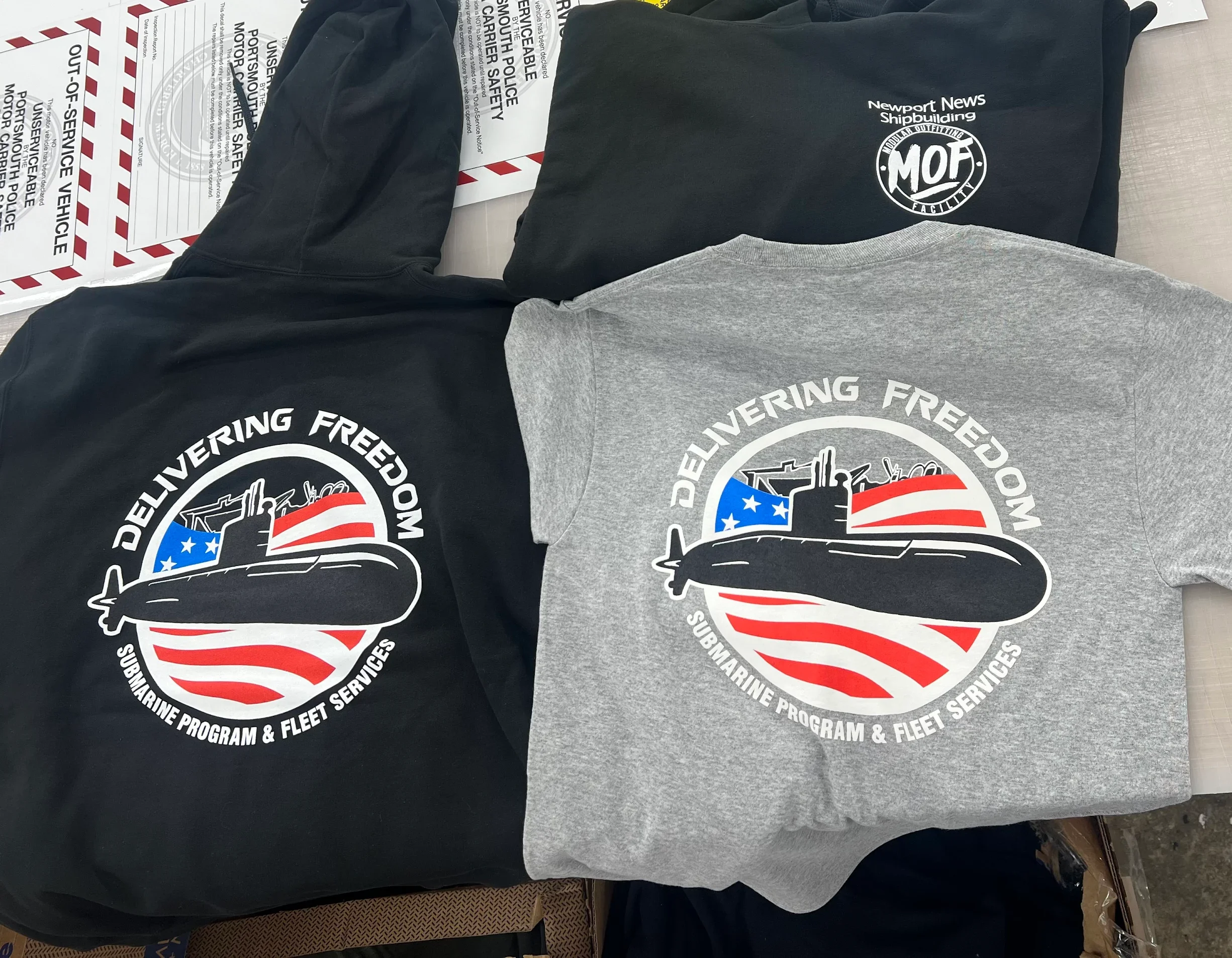 Two sweatshirts with a navy submarine graphic and text 'Delivering Freedom' and 'Submarine Program & Fleet Services' in front of some government postal service envelopes.