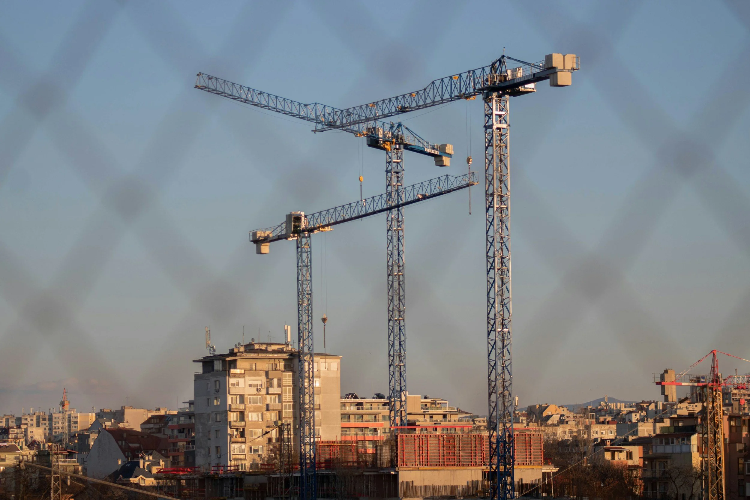 Three blue construction cranes over a cityscape with buildings, under a partly cloudy sky.