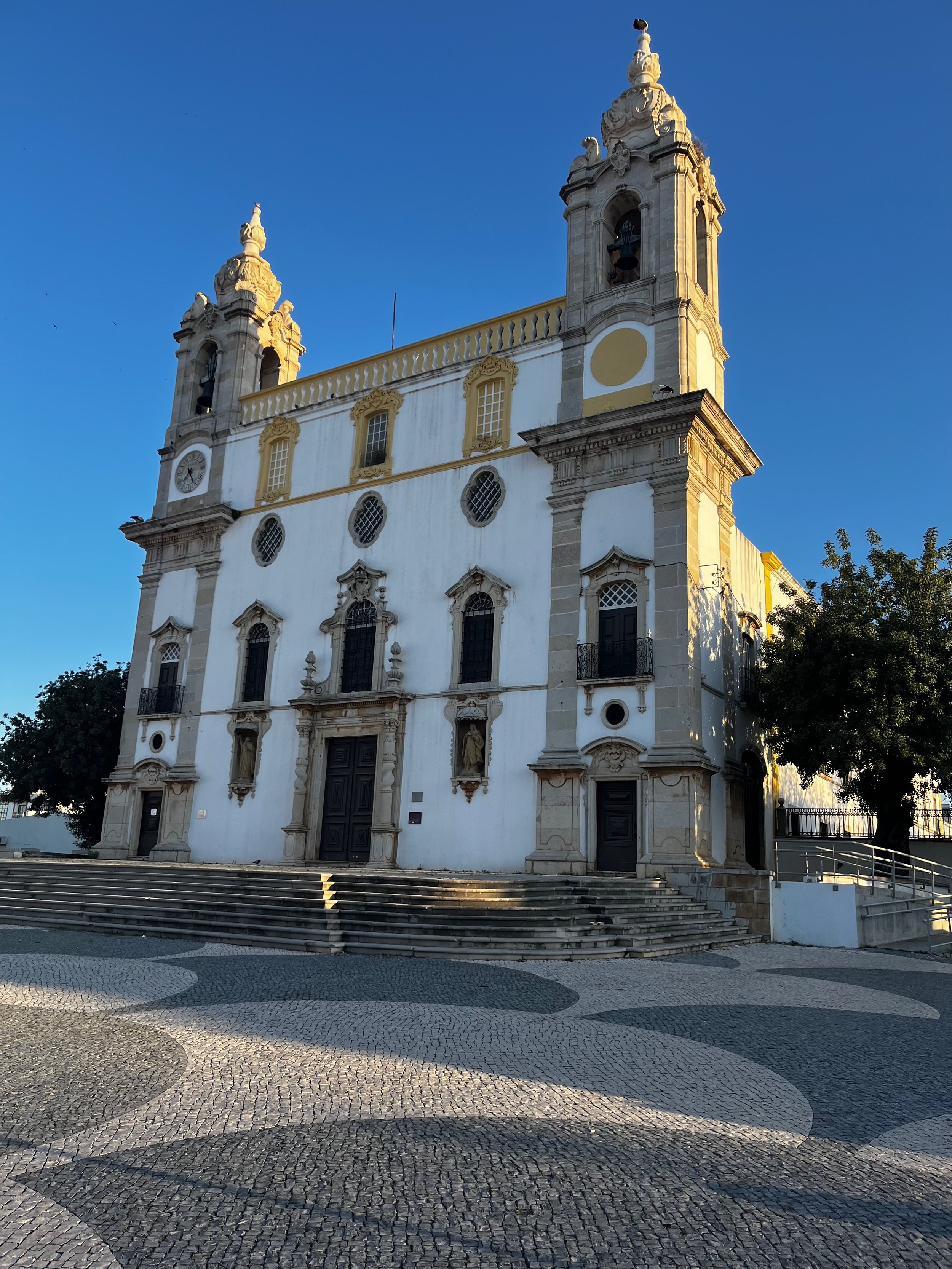 A historic white and yellow baroque-style church with two bell towers under a clear blue sky, featuring ornate window frames, statues, and decorative details, with steps leading up to the entrance and trees nearby.