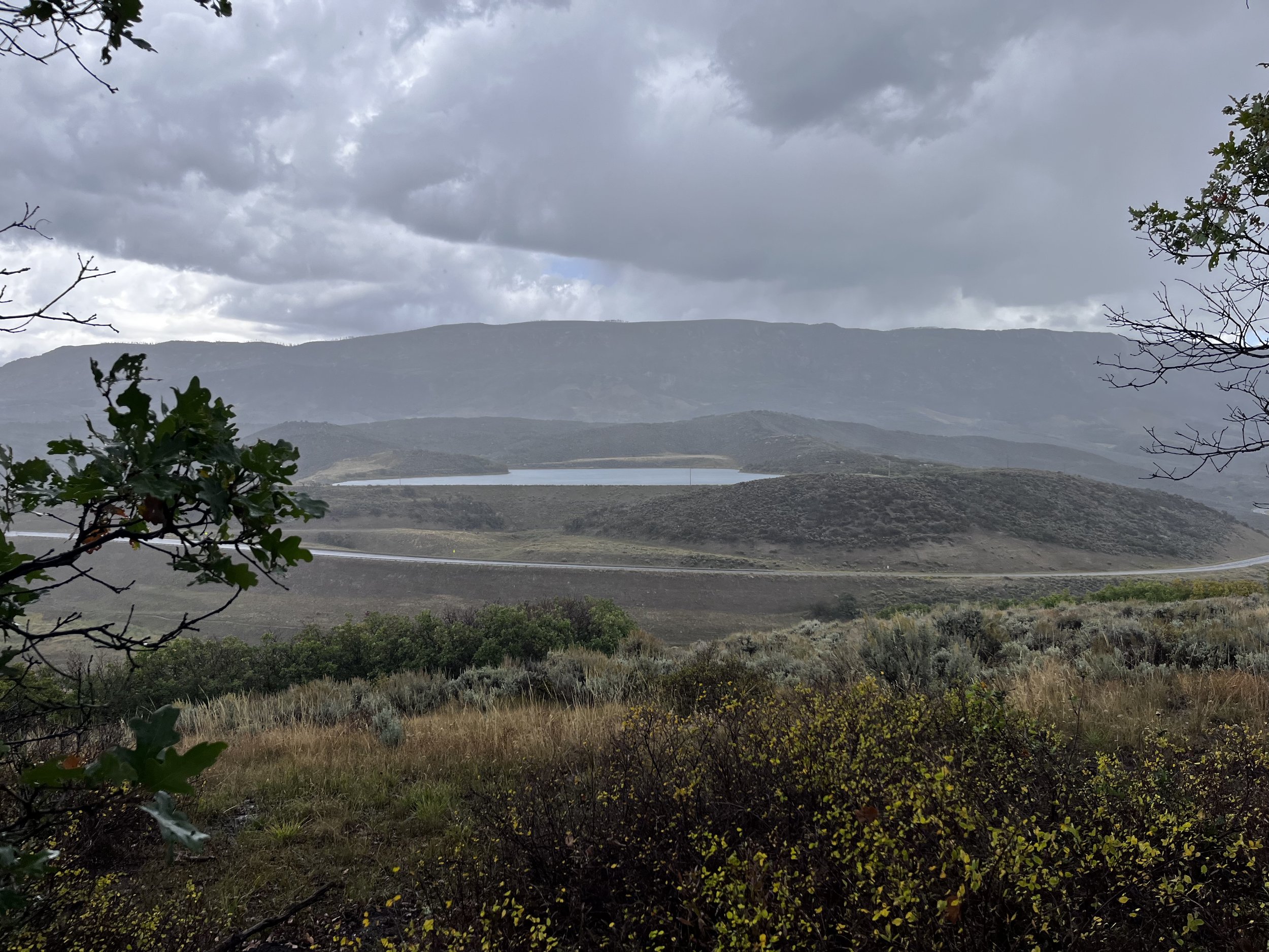 Rain clouds over rolling hills, a reservoir, and a winding road viewed from Cerro Summit, Colorado.