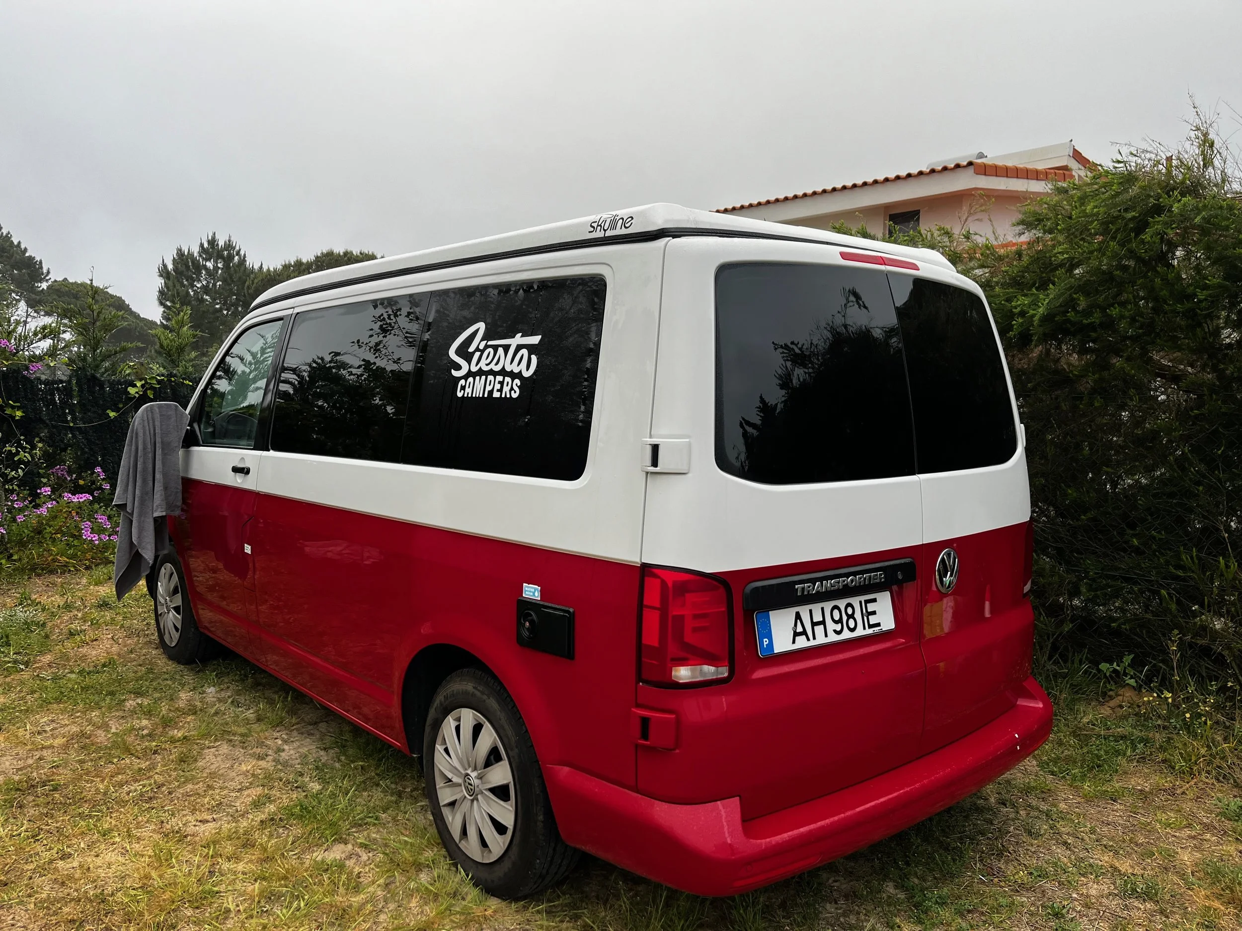 A red and white Volkswagen camper van parked on a grassy area with trees and a house in the background, and a gray towel hanging from the driver's side mirror. The van has a sticker that says 'Siesta Campers' on the window.