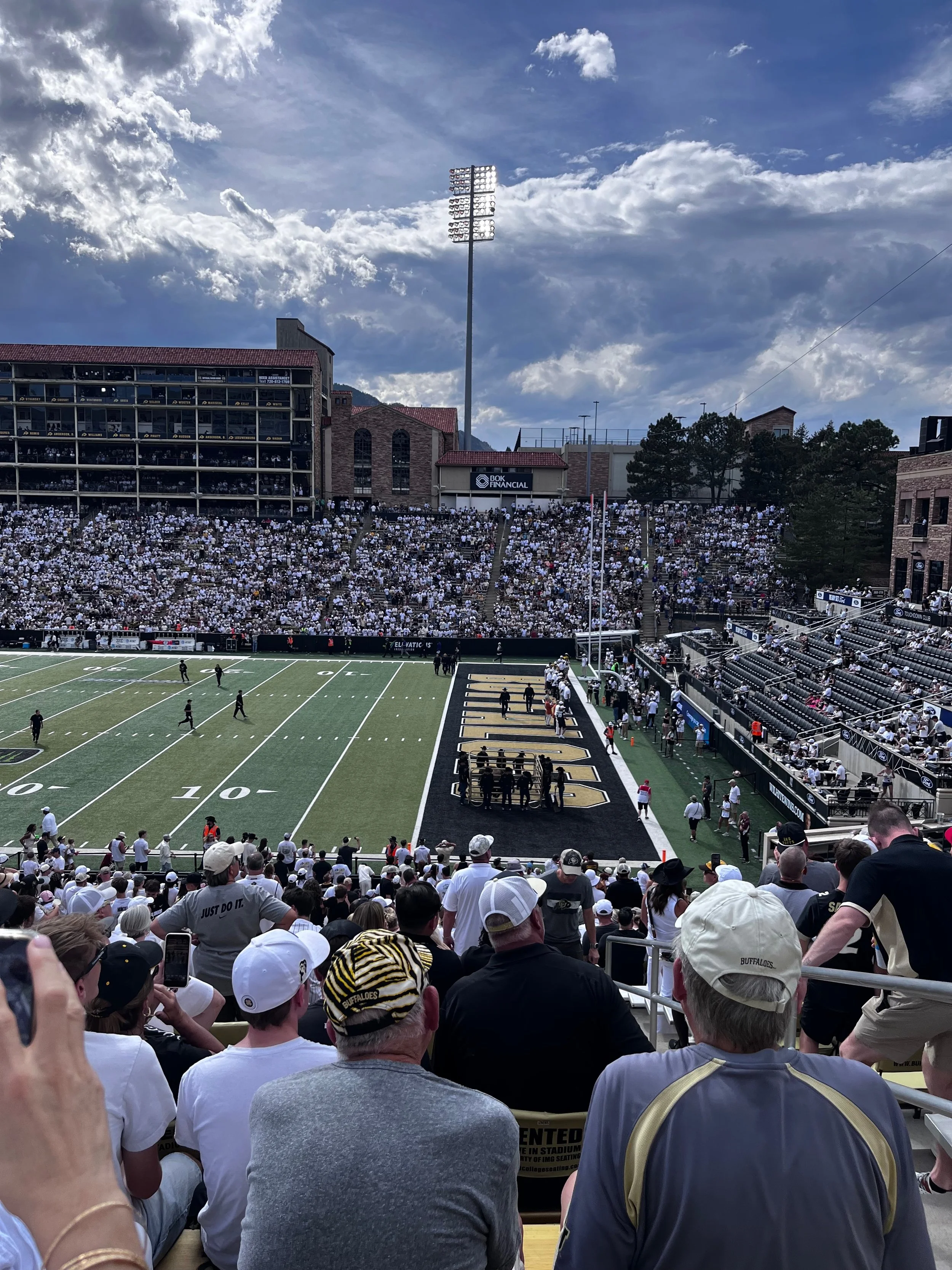 View from the stands over the Colorado Buffaloes end zone at Folsom Field, with fans filling the opposite stands and Balch Field House beyond them.