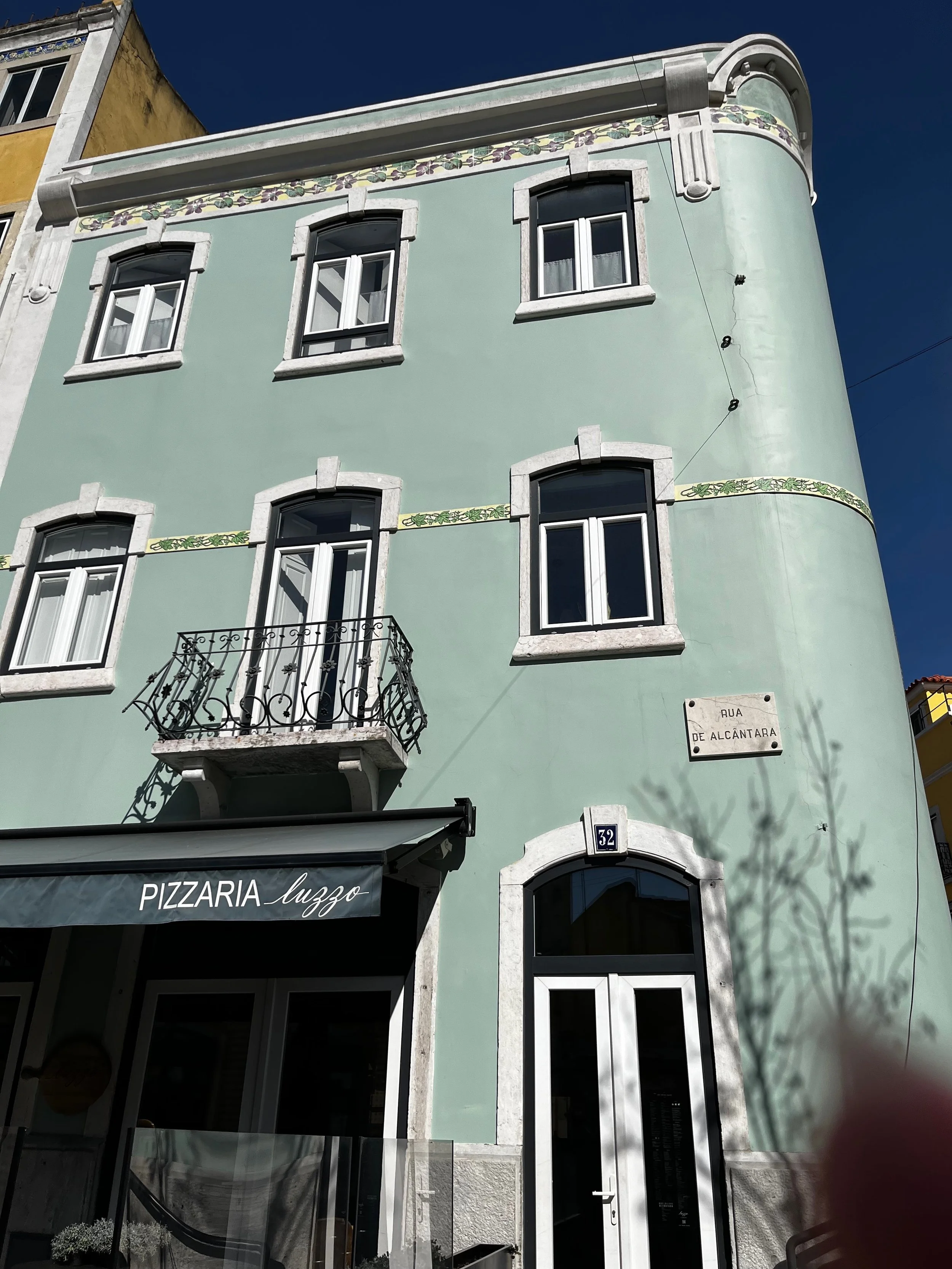 Rounded pale green Art Deco-style building with decorative tile and wrought-iron balcony in Alcântara, Lisbon.