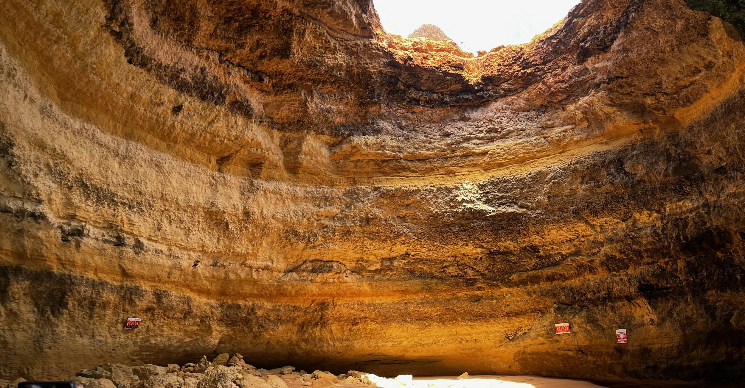 Large cave with a wide, rocky ceiling and a bright opening at the top, illuminated by sunlight, with warning signs on the walls and a rocky floor.