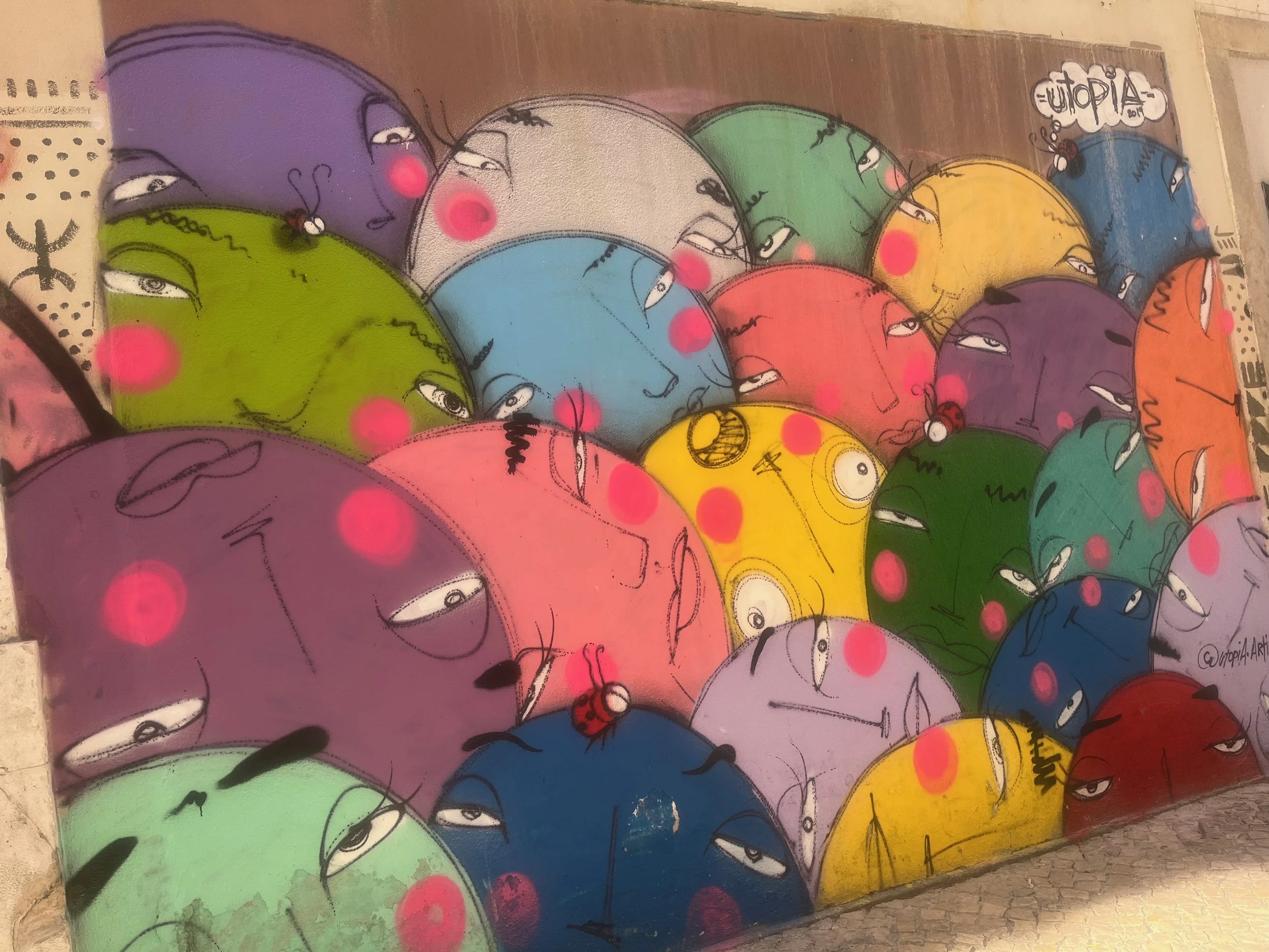 Colorful graffiti street art featuring overlapping cartoonish faces in various bright colors with pink blush spots.