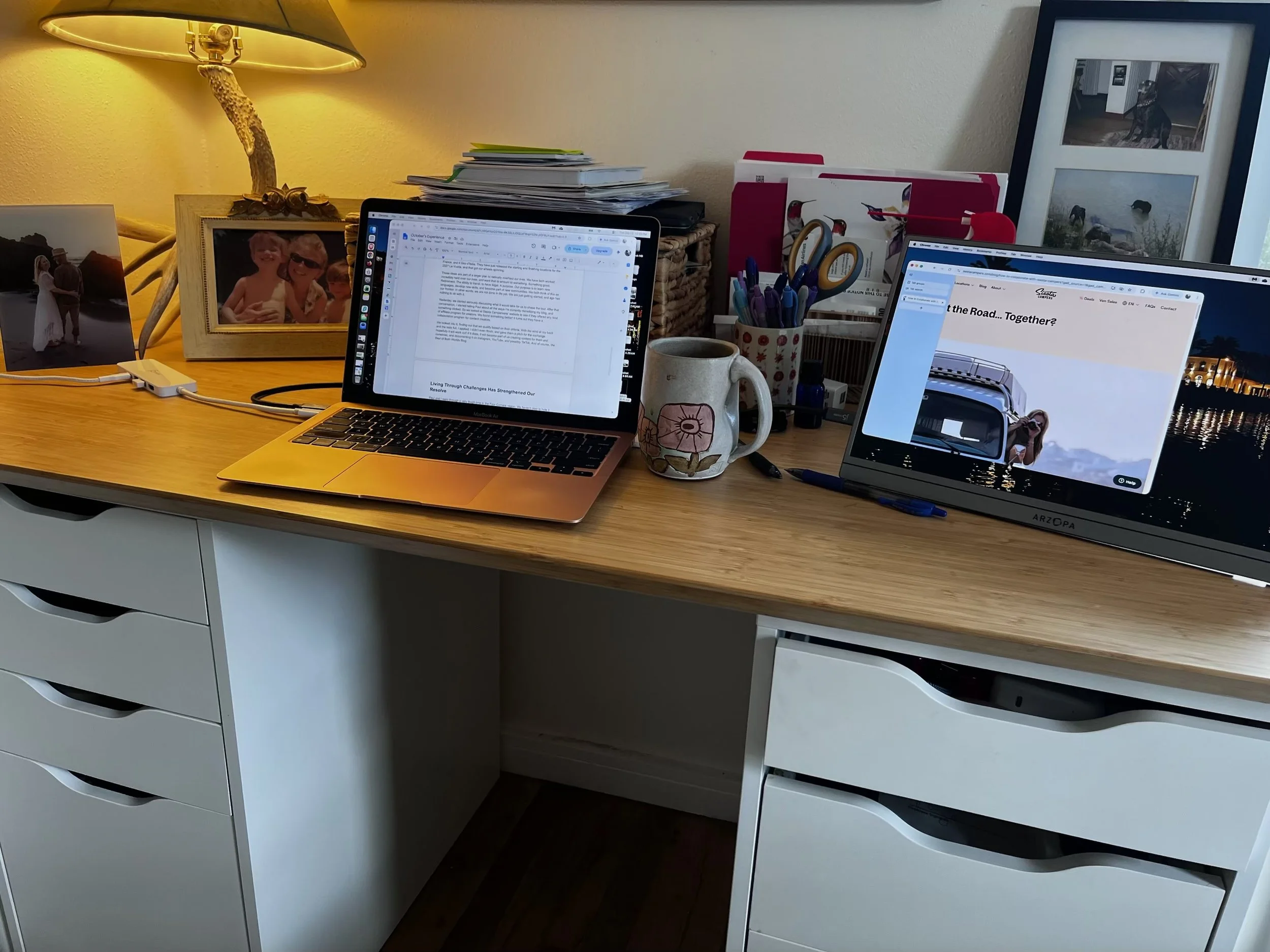 A cluttered desk with two laptops, a coffee mug, pens, and framed photos, illuminated by a yellow lamp, with organized stacks of papers and folders behind.