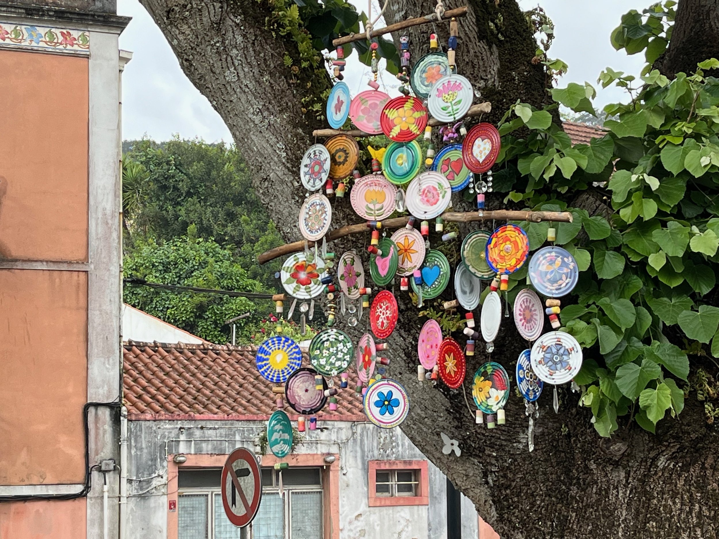 Colorful decorative wind chimes hanging from a large tree against a backdrop of buildings and a cloudy sky.