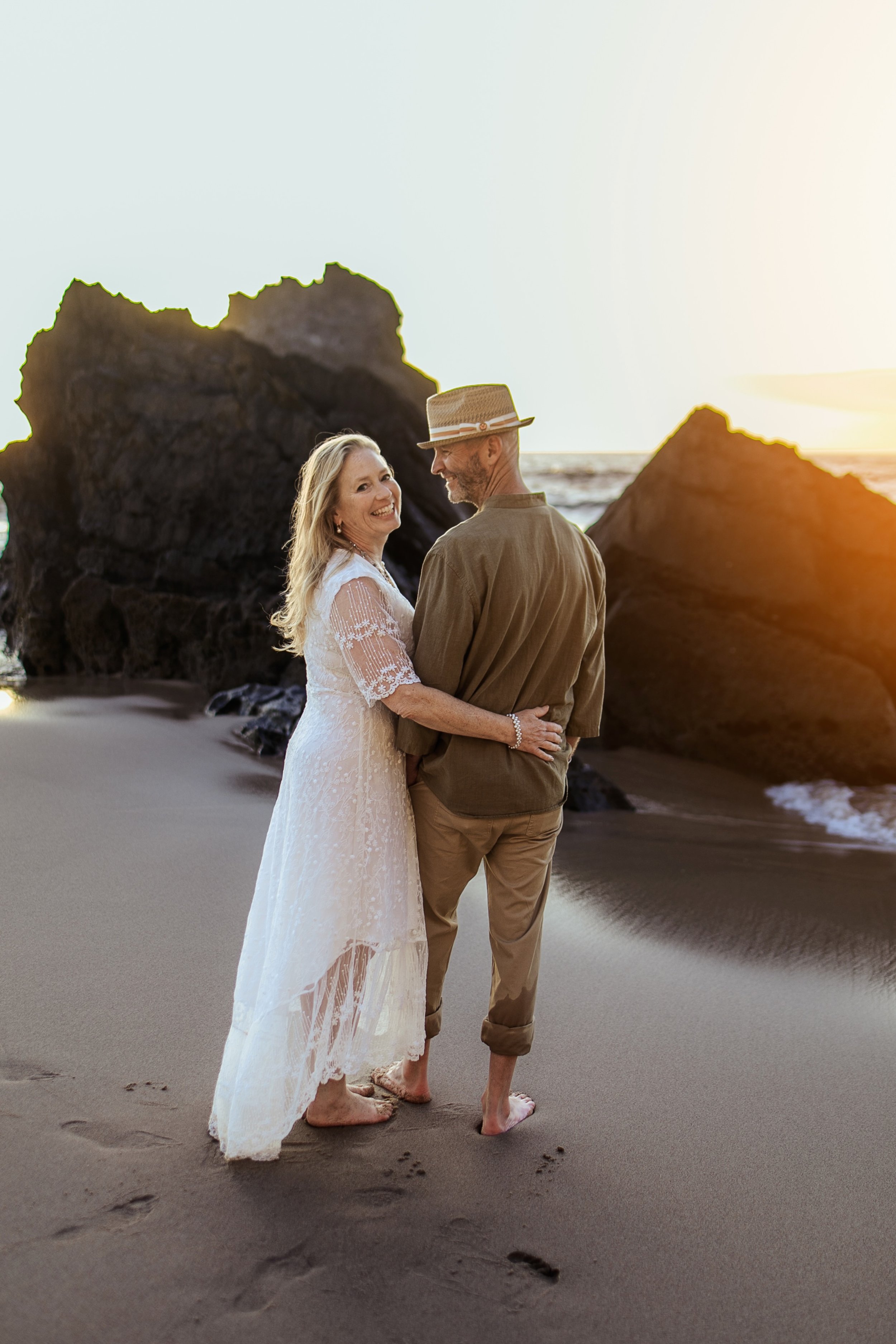 A smiling woman in a white lace dress and a man in a tan shirt and pants, both barefoot, standing together on a sandy beach near large rocks during sunset.