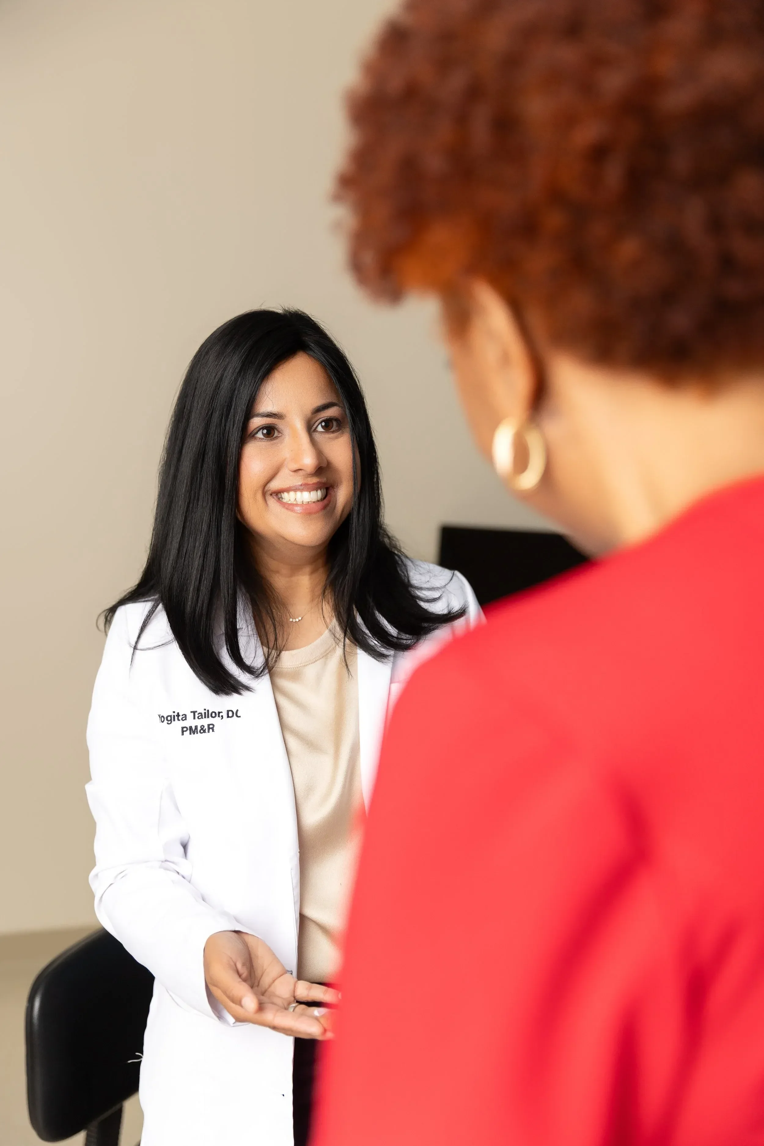 Dr. Yogita Tailor with long black hair and a white lab coat smiling and looking at a person in a red top, with their back facing the camera.