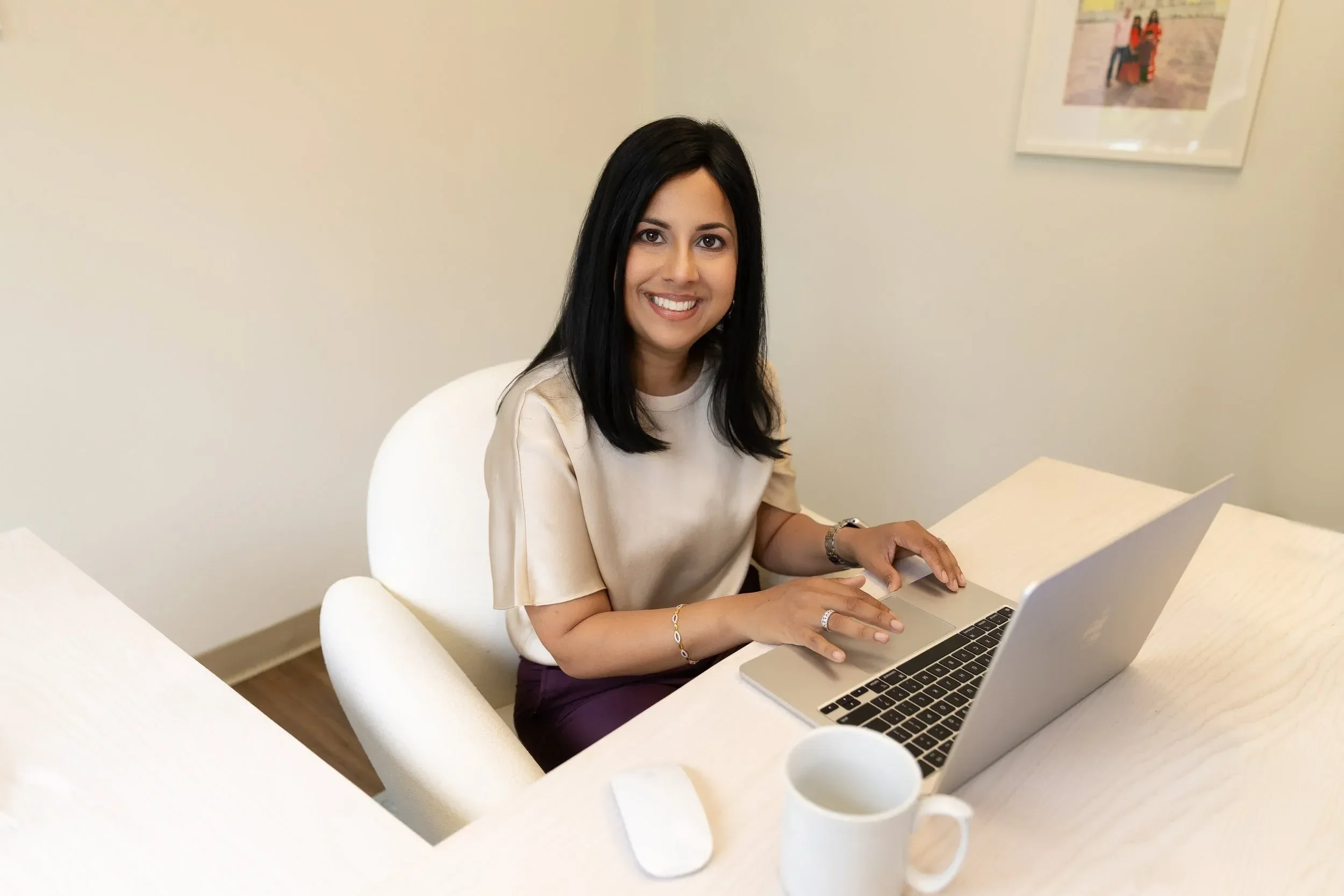 Dr. Yogita Tailor with black hair and a beige shirt sitting at a white table, smiling, working on a silver laptop, with white mugs on the table, in a room with white walls and a colorful picture hanging on the wall.