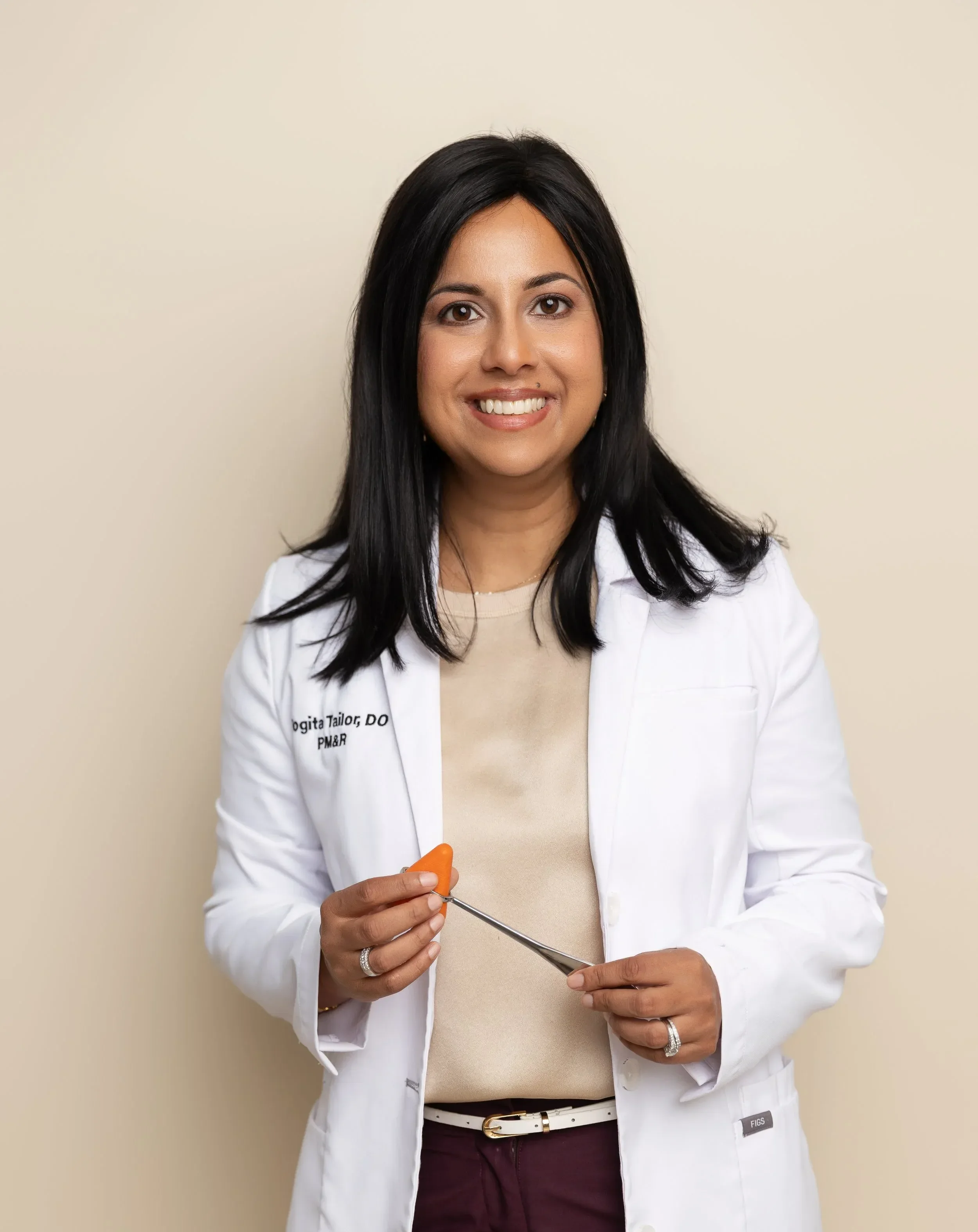 Dr. Yogita Tailor with dark hair wearing a white medical coat and beige top, standing against a beige wall, holding a surgical instrument.