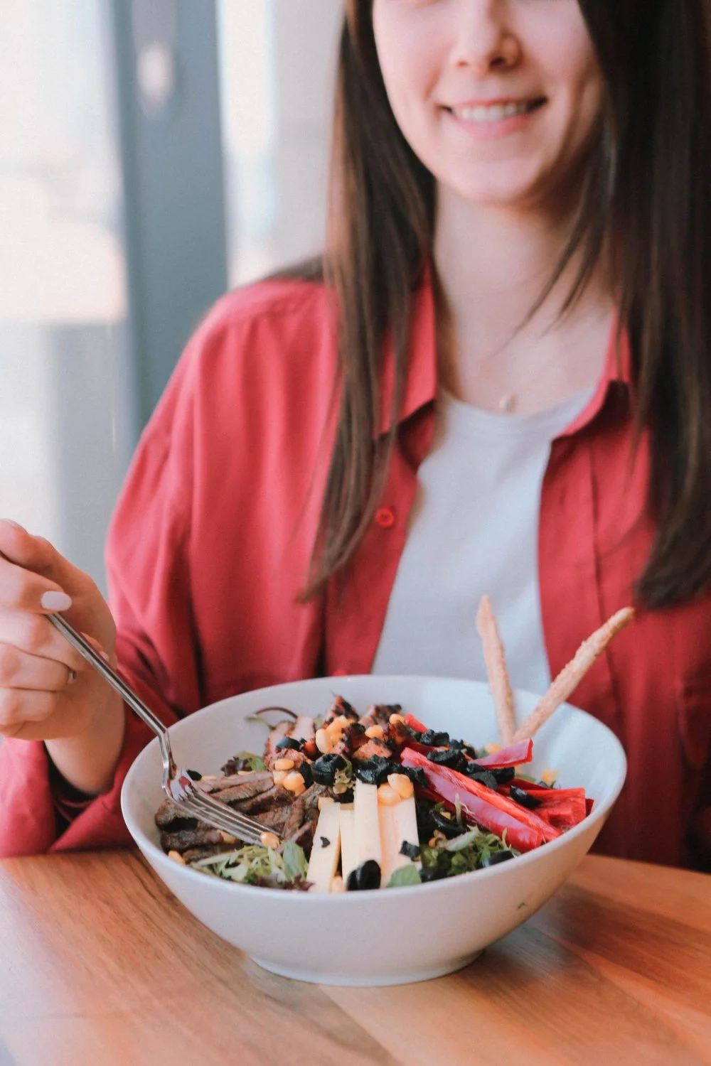 A woman in a coral shirt and white top sitting at a table with a bowl of salad, holding a fork, smiling.