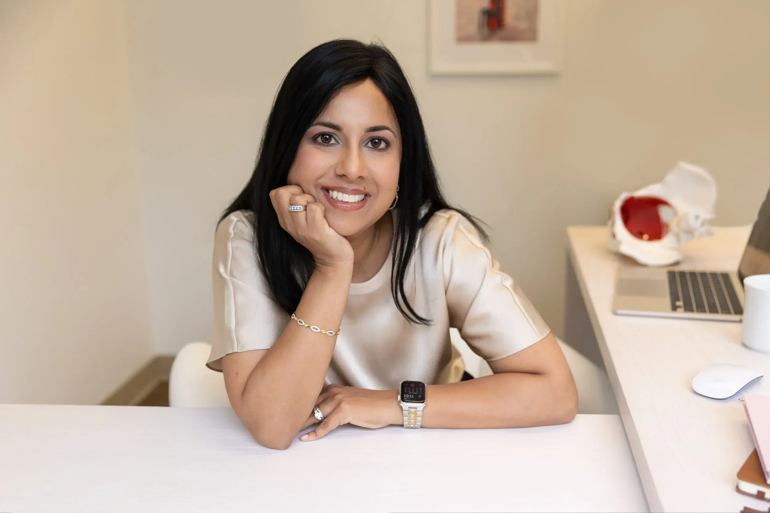 Dr. Yogita Tailor with long black hair and brown eyes sitting at a white desk, resting her chin on her hand, smiling, wearing a beige top, a gold bracelet, rings, and a smartwatch, with a model of a skull and a laptop on her right.