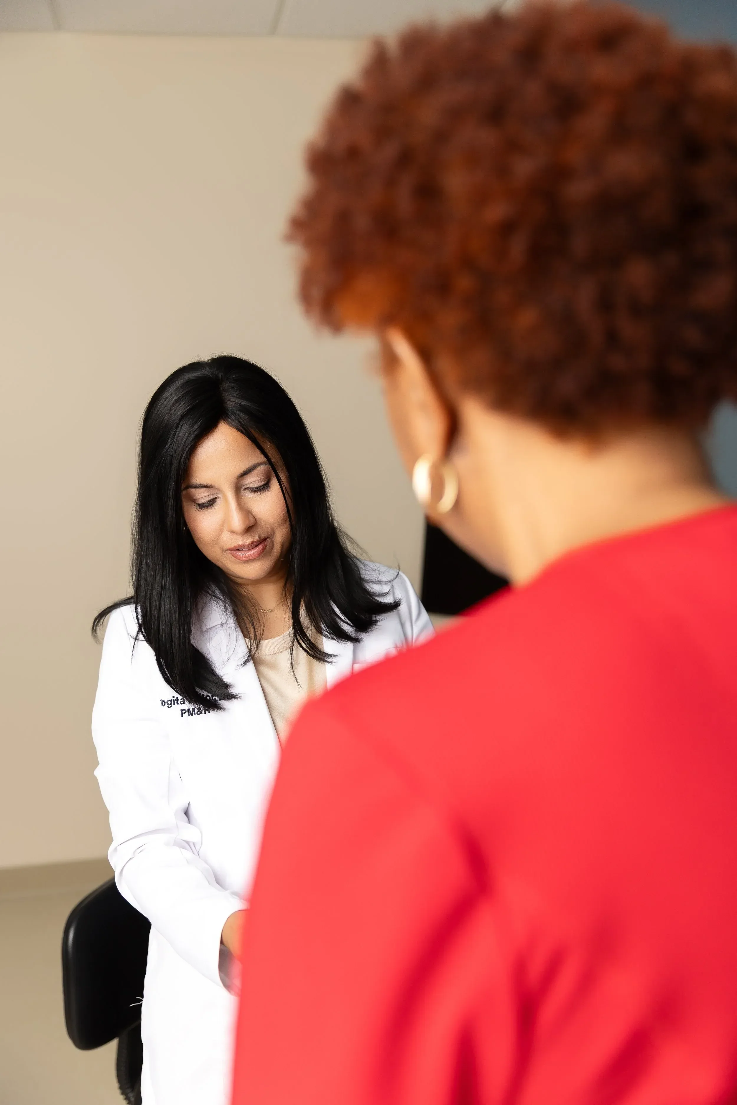 A nurse with long black hair wearing a white lab coat looking down, and a person with short curly auburn hair and earrings facing away from the camera in a red shirt.