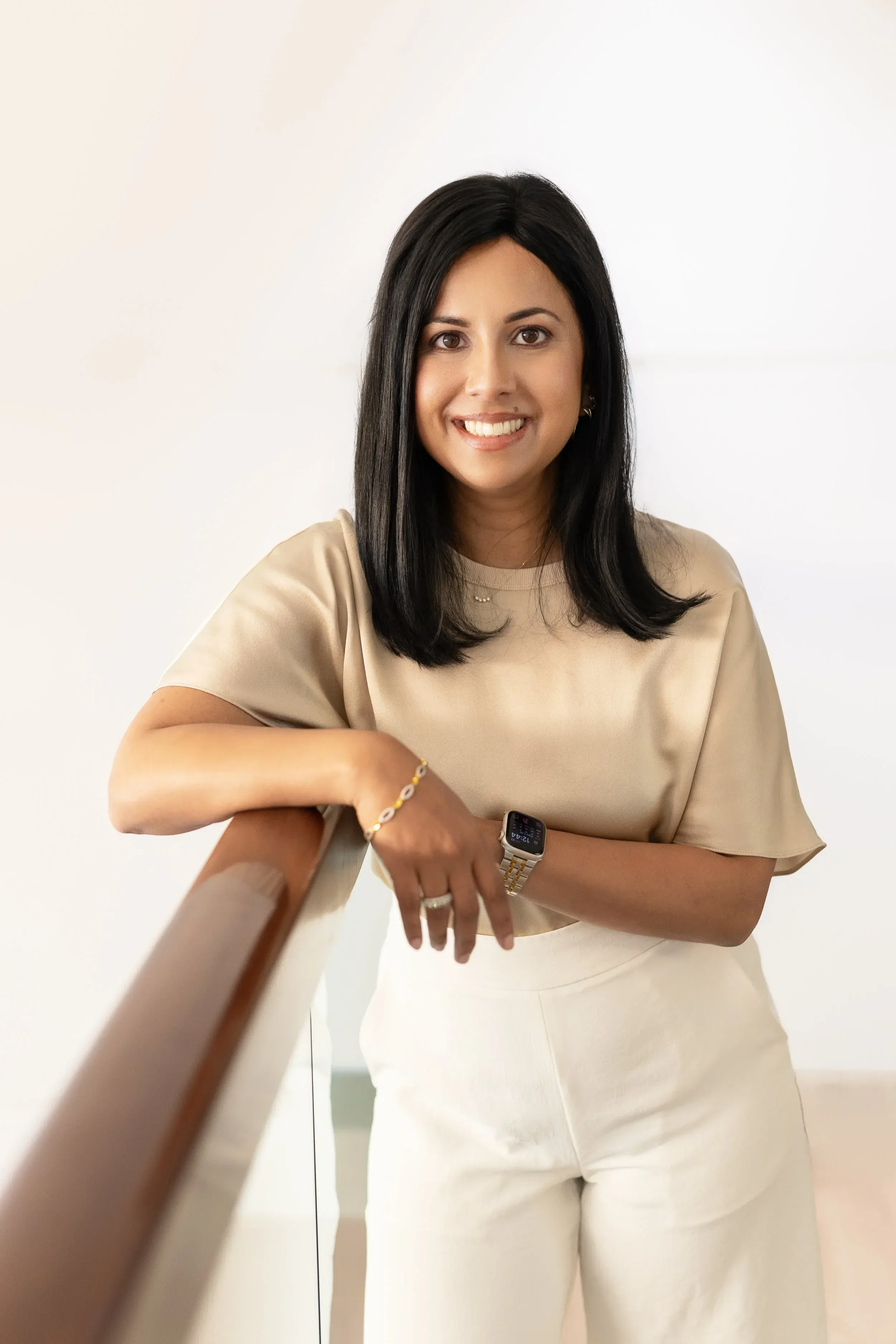 Dr. Yogita Tailorn with black hair smiling, wearing a beige blouse, white pants, a bracelet, and a watch, leaning on a wooden railing in front of a white wall.